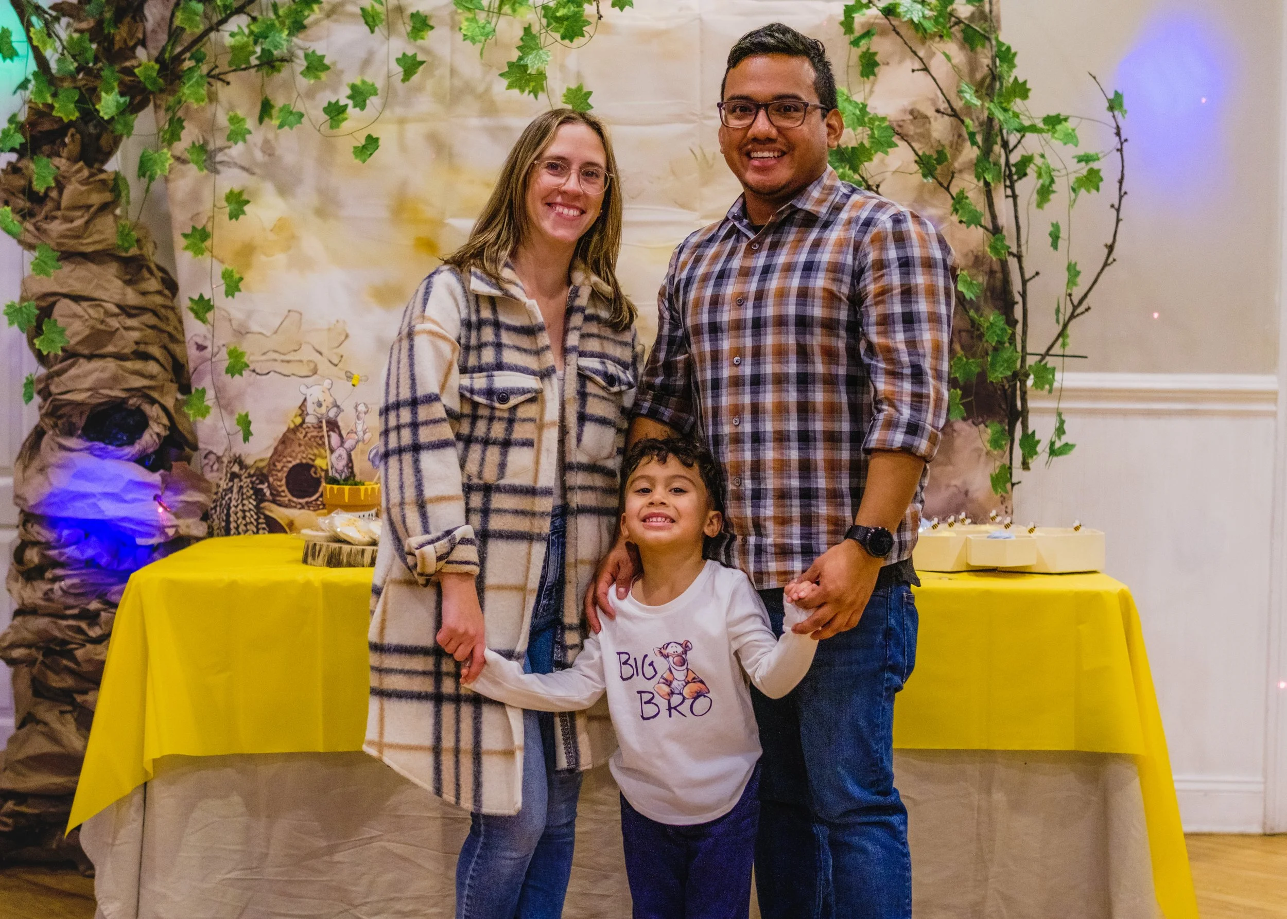 A family of three smiling and holding hands in front of a table with a children's birthday party theme backdrop with a lion and tree. The woman is wearing a plaid coat, the man is in a plaid shirt, and the child is in a white shirt with a tiger and t