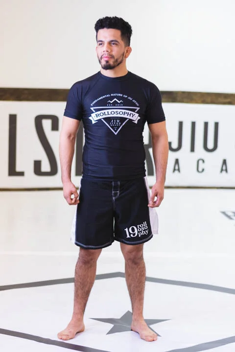 Man standing on a gym mat at Jiu Jitsu academy, wearing a black T-shirt and black shorts, with a serious expression.