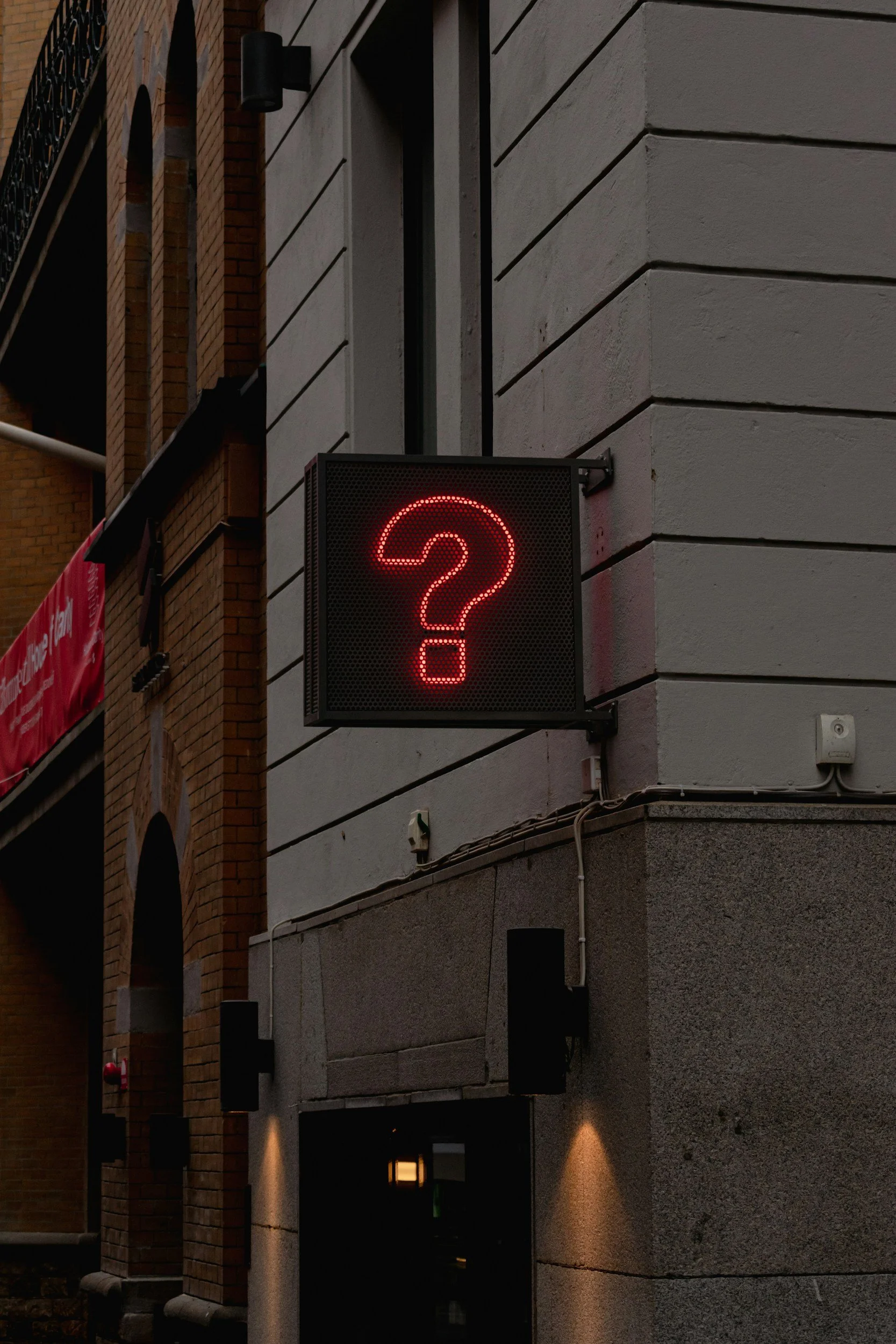 A street scene showing a building with a gray wall and a red digital question mark sign mounted on the wall.