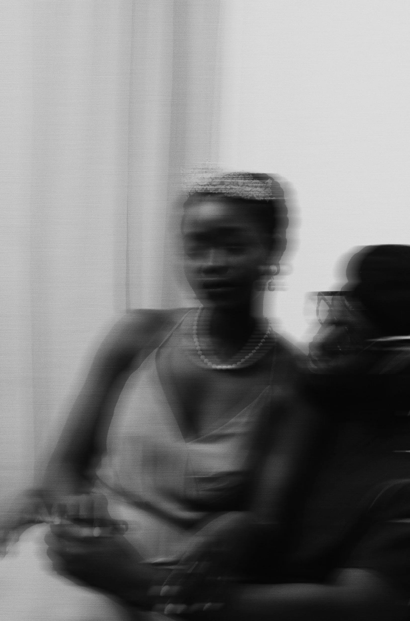 Black and white photo of a woman taking a mirror selfie with a smartphone, wearing earrings, a necklace, and a sleeveless top, in front of a plain background.
