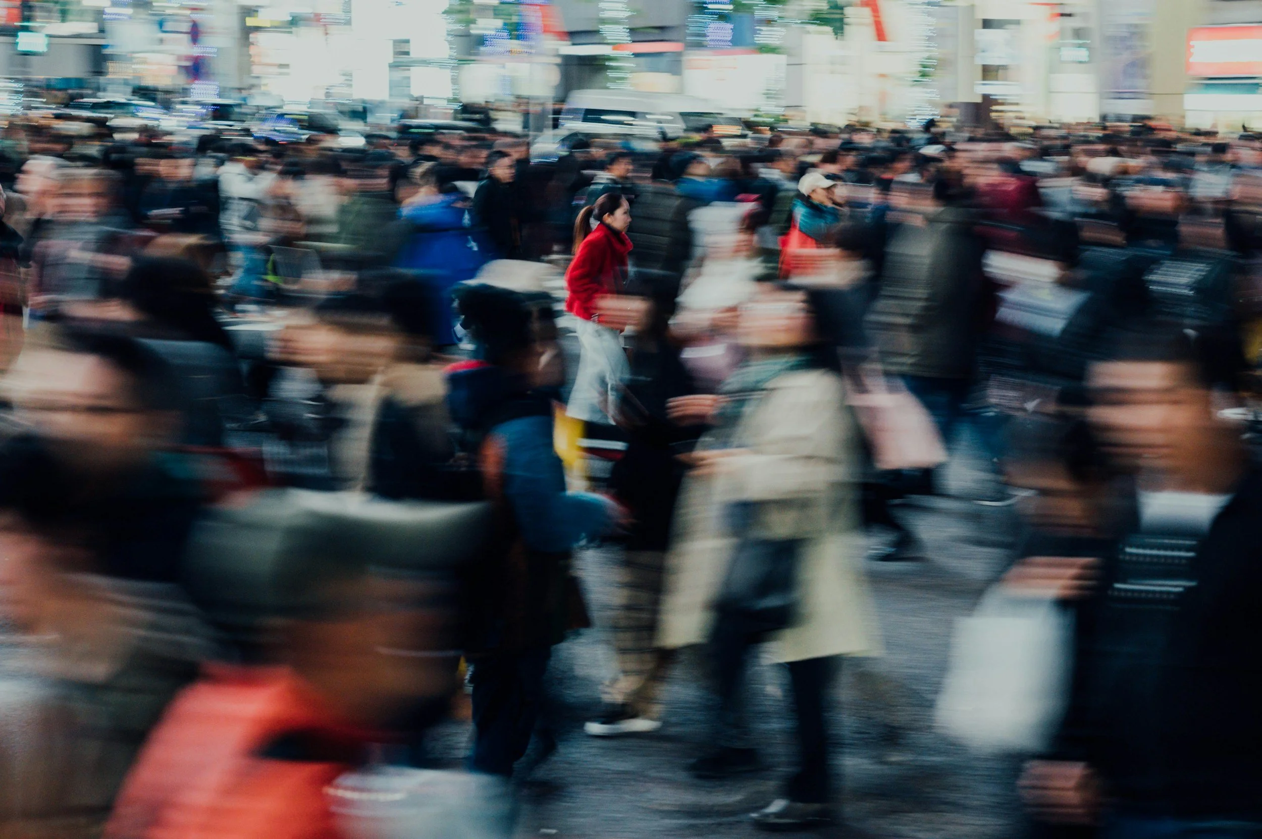 A crowded city street scene with many blurred pedestrians walking, some on motorcycles and scooters, creating a sense of motion.