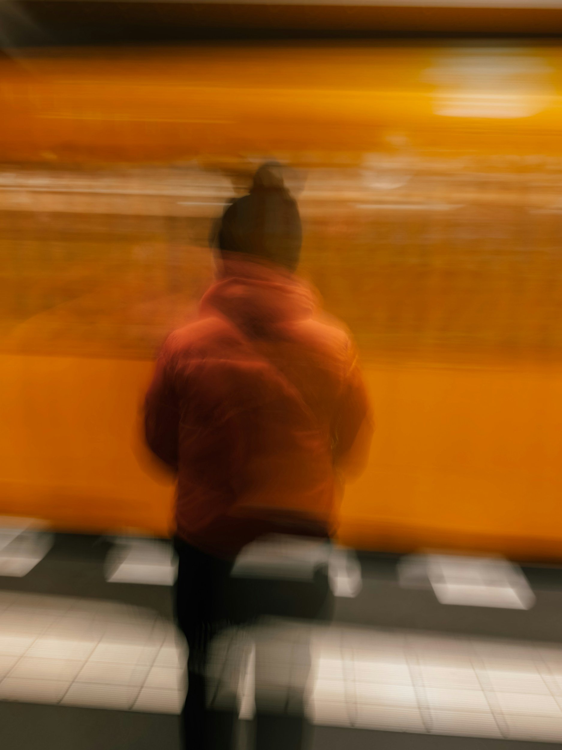 A blurry photo of a person taking a selfie in a mirror. The person is wearing a dark jacket and pants, and has long hair tied back. The background includes an orange wall and a tile floor.