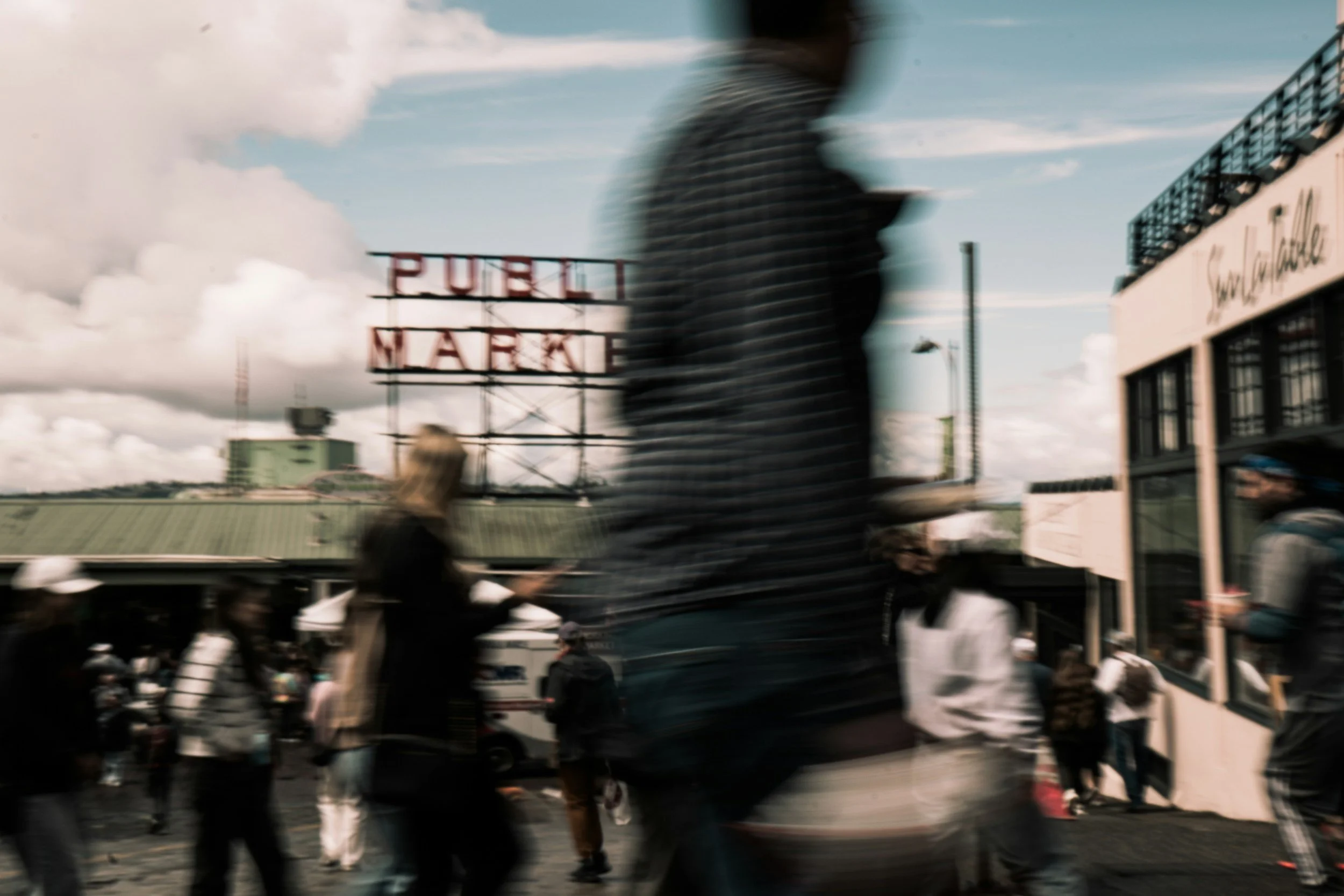 A crowded city street scene with numerous blurred pedestrians walking past, in front of a building with a sign that reads 'PUBLIX MARKET', under a blue sky with scattered clouds.