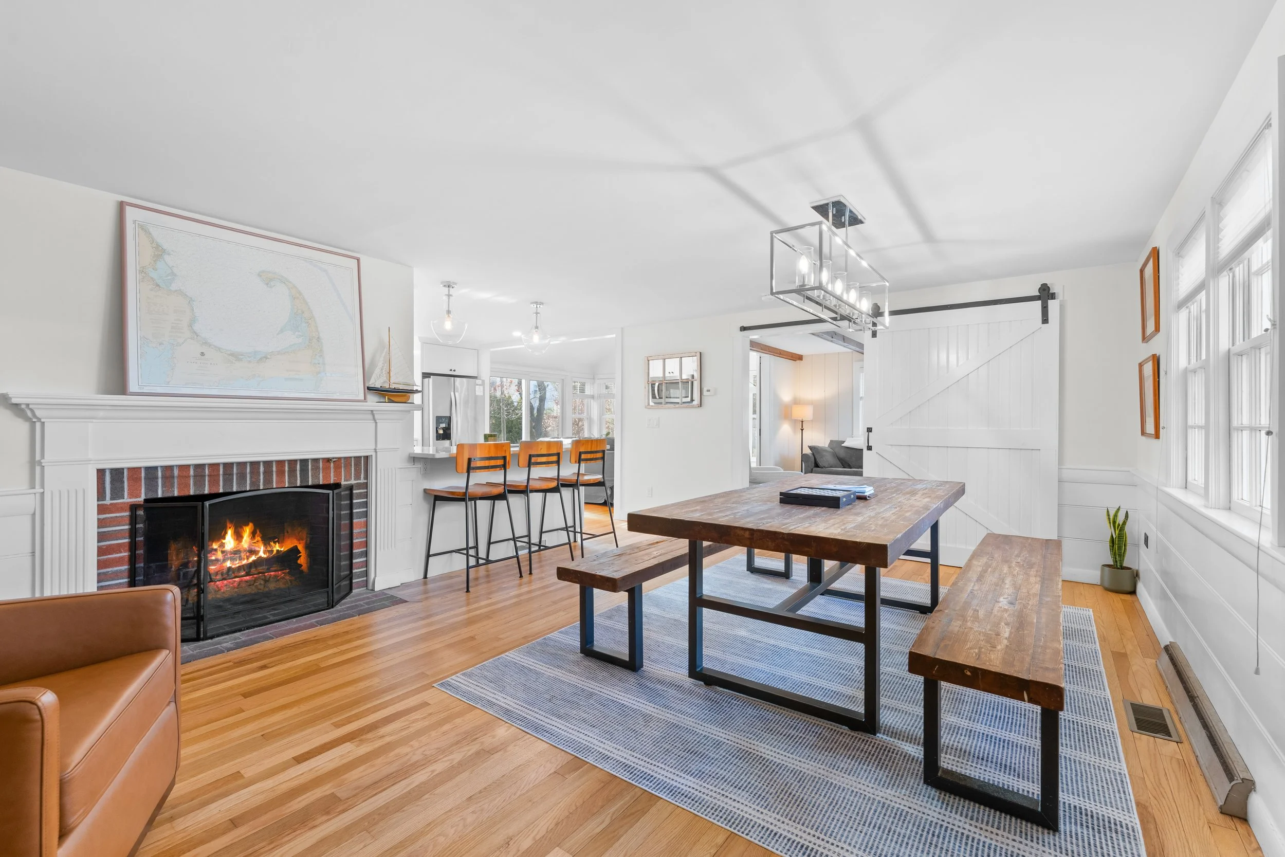Bright living room with a brick fireplace, white walls, large windows, wooden furniture, and modern light fixtures.