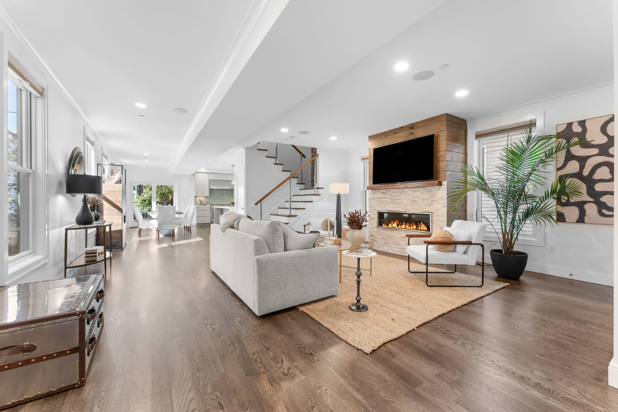 Spacious modern living room with white walls and dark hardwood floors. Features a white sofa, armchair, large potted plant, flat-screen TV above a fireplace with stone surround, and a wooden accent wall around the TV. Adjacent to a dining area and ki