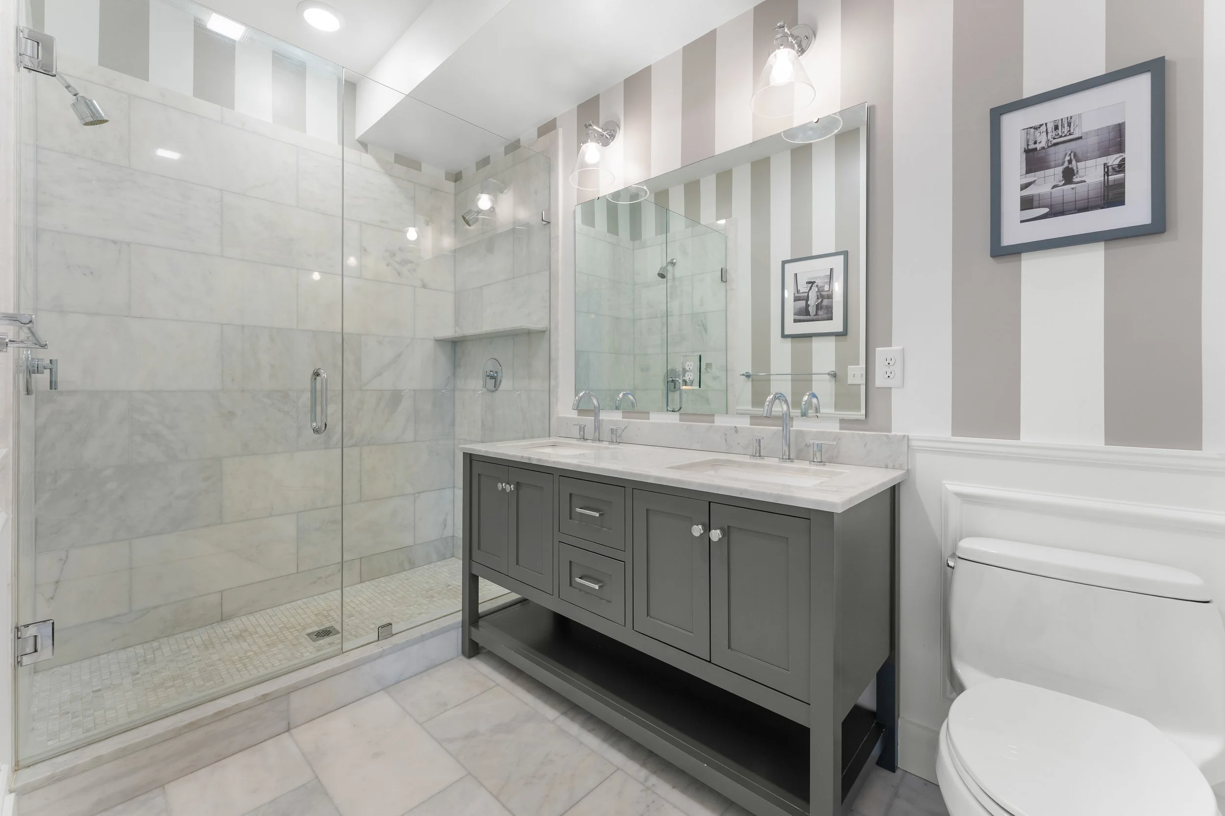 Modern bathroom with marble shower, double sink vanity, framed black-and-white photographs on striped wall, and a toilet.