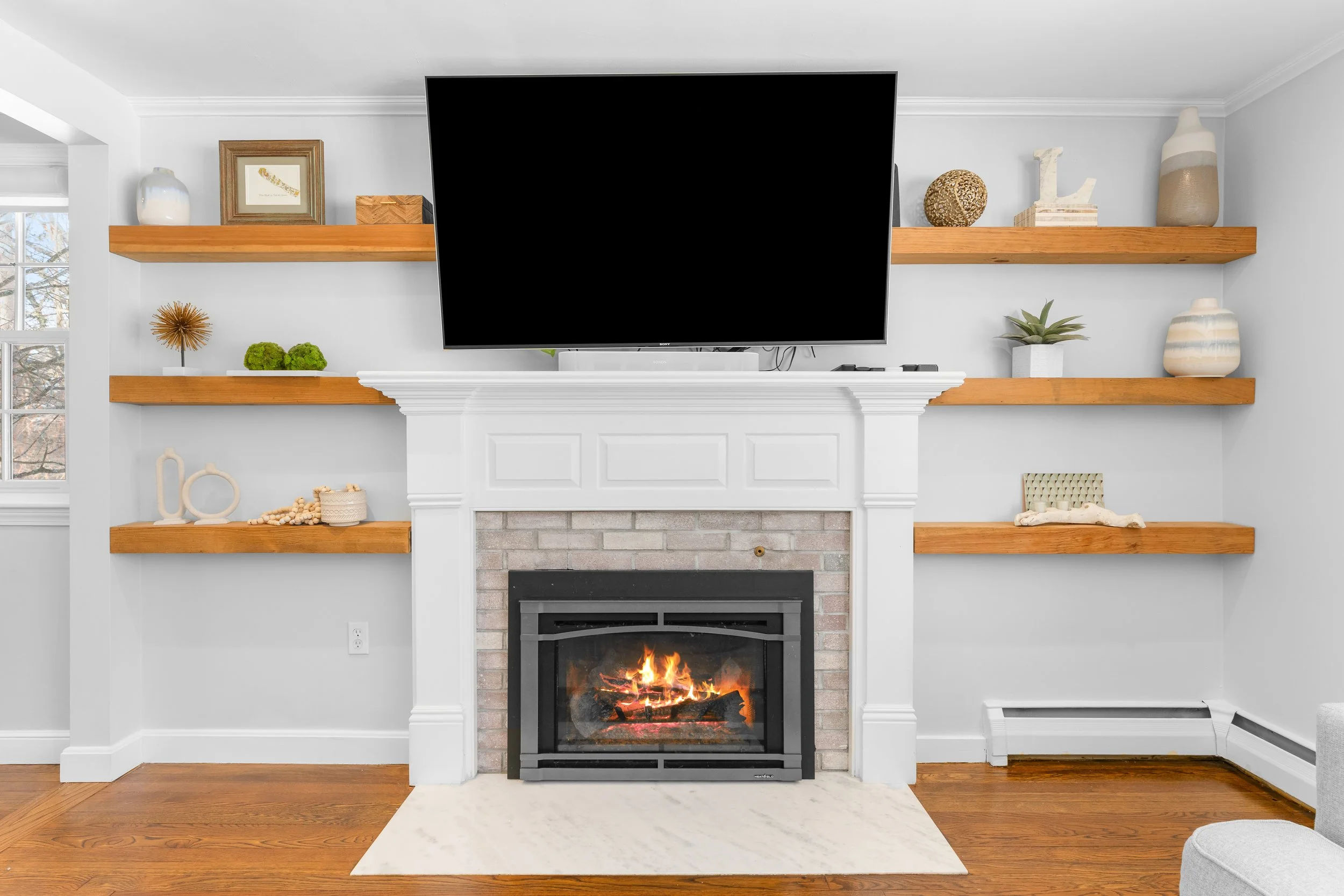Living room with a fireplace and a flat-screen TV mounted above it, decorated with wooden shelves holding various decorative objects and plants.