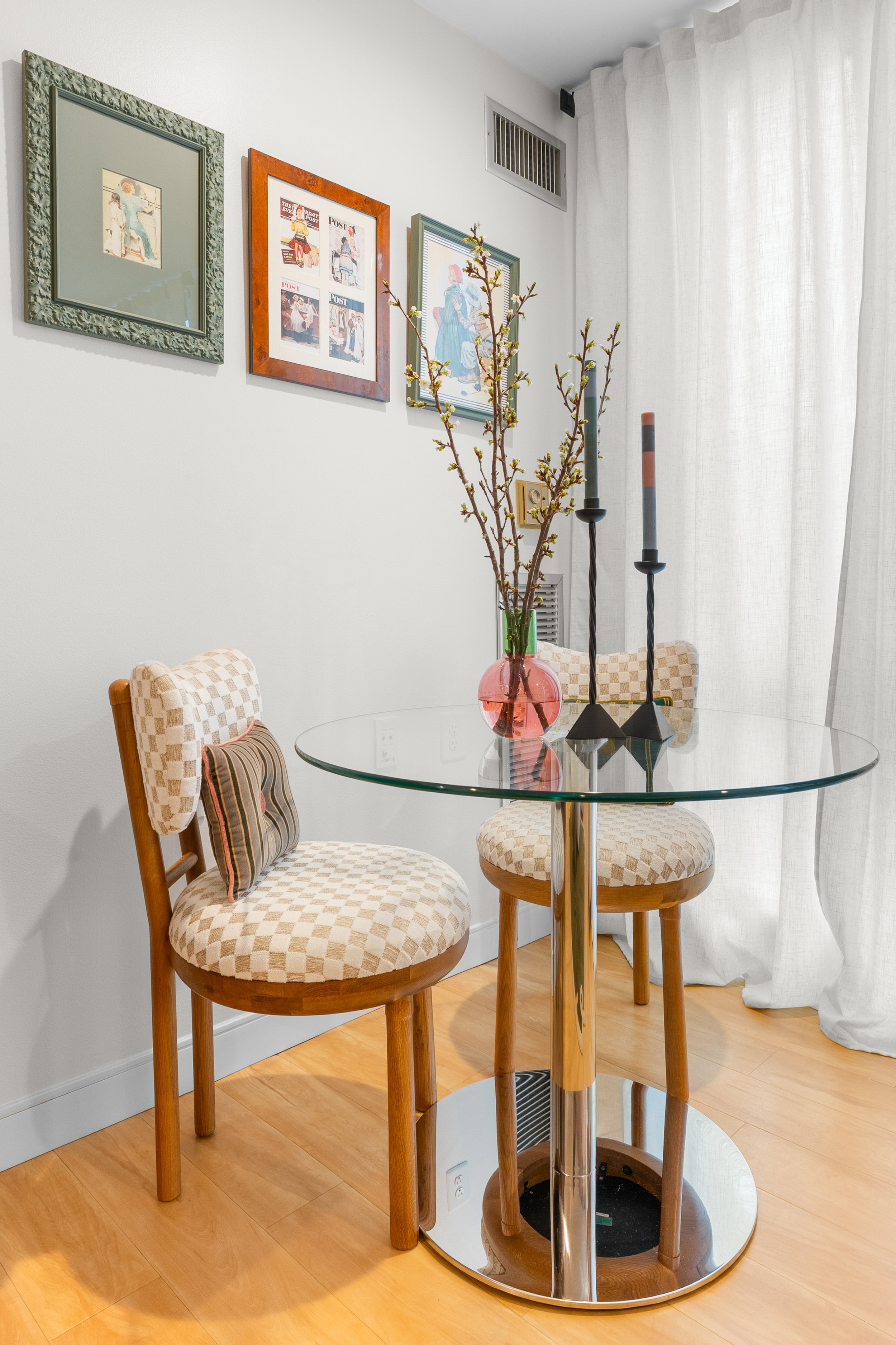 A glass-top round dining table with two wooden chairs with padded checkered beige and white cushions and striped pillows, a pink vase with blossoming branches, and two tall black candlesticks inside a bright, minimal kitchen or dining area with wall art, white curtains, and hardwood flooring.