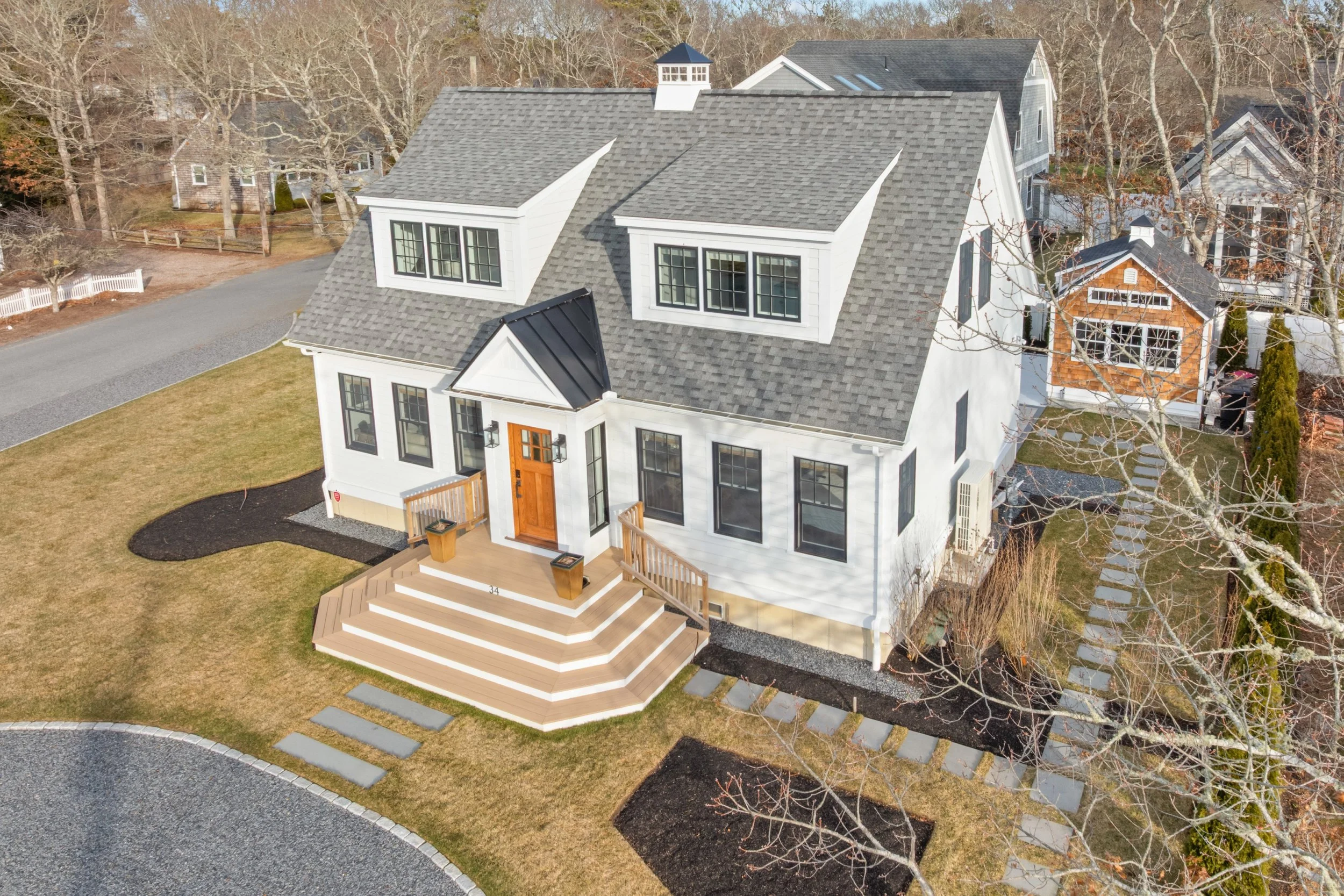 Aerial view of a modern white house with large windows, a gray shingle roof, and a wooden front porch with steps. The house is surrounded by a lawn, pathways, and neighboring houses in a wooded area with leafless trees.