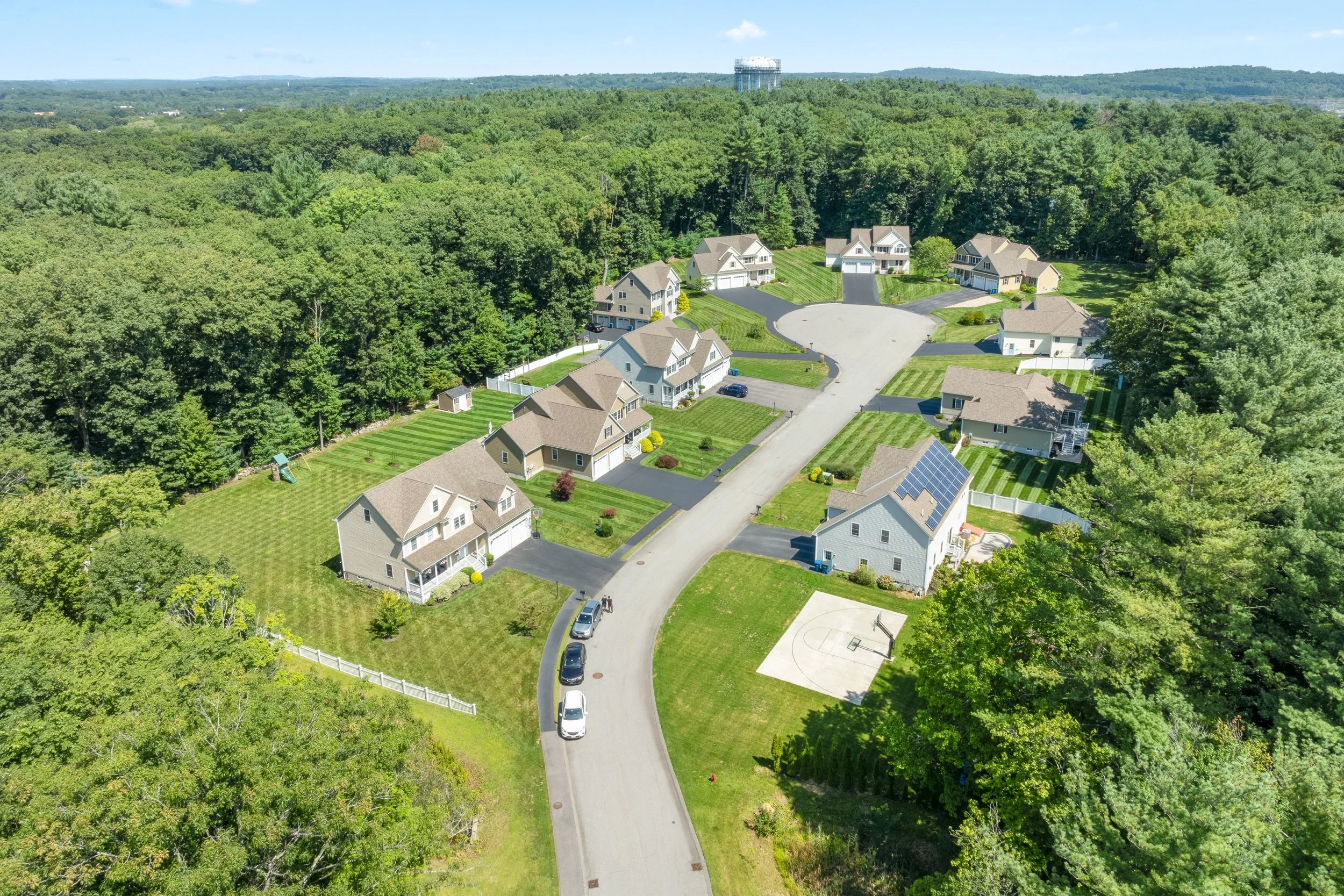 Aerial view of a suburban neighborhood with multiple houses, well-maintained lawns, and a basketball court, surrounded by a dense tree line in the background.