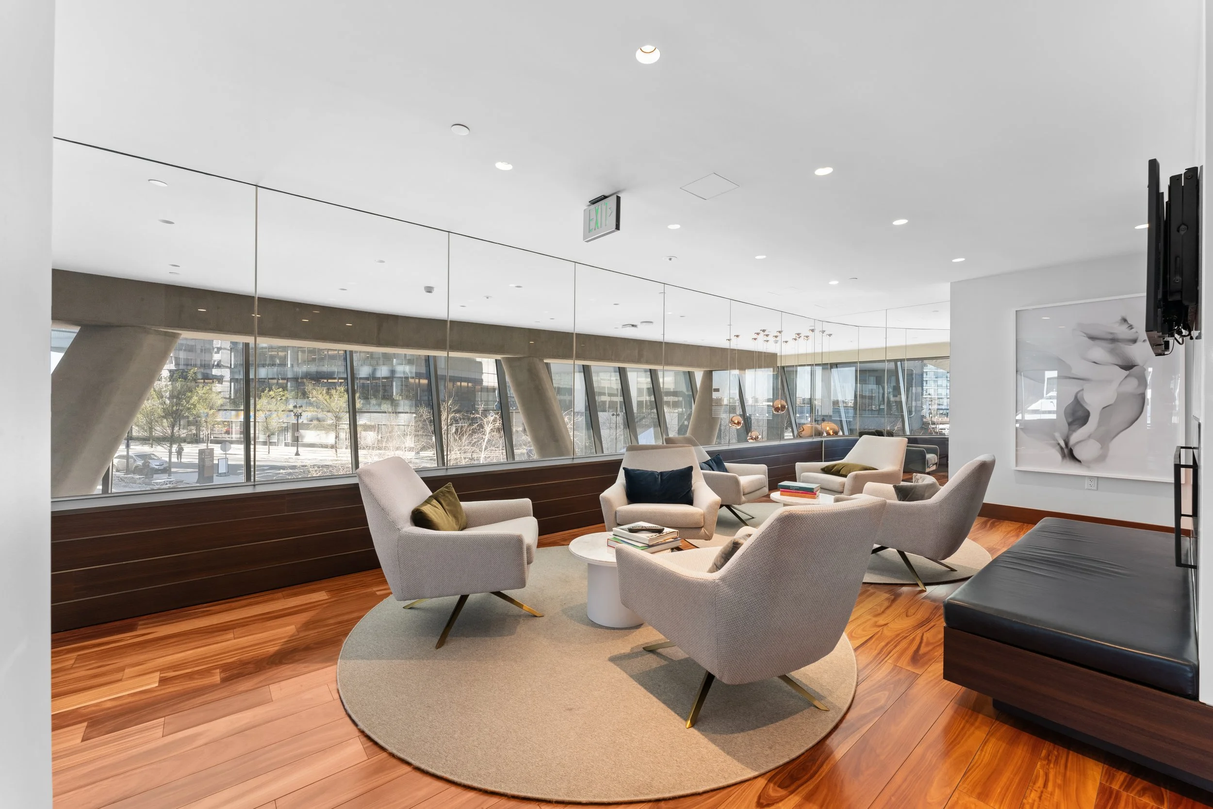 Modern indoor lounge area with white armchairs around a small round coffee table, large windows revealing a cityscape, and contemporary artwork on the wall.
