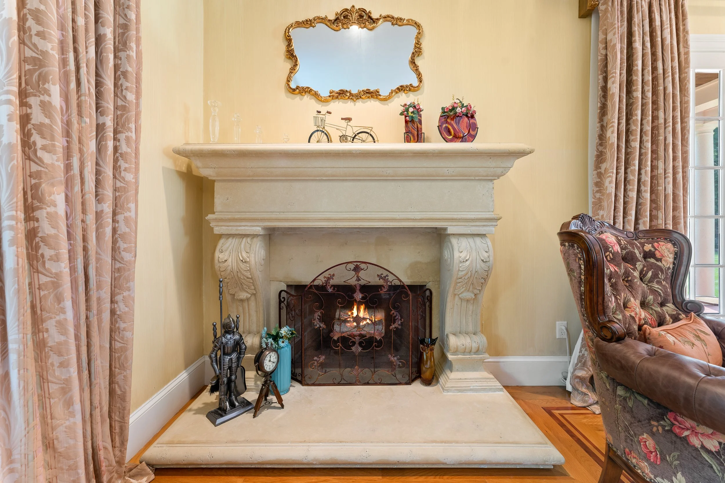 A decorated living room corner with a fireplace, a mirror above it, and various decorative items on the mantel. To the left are pink floral curtains, and part of a floral armchair is visible on the right.