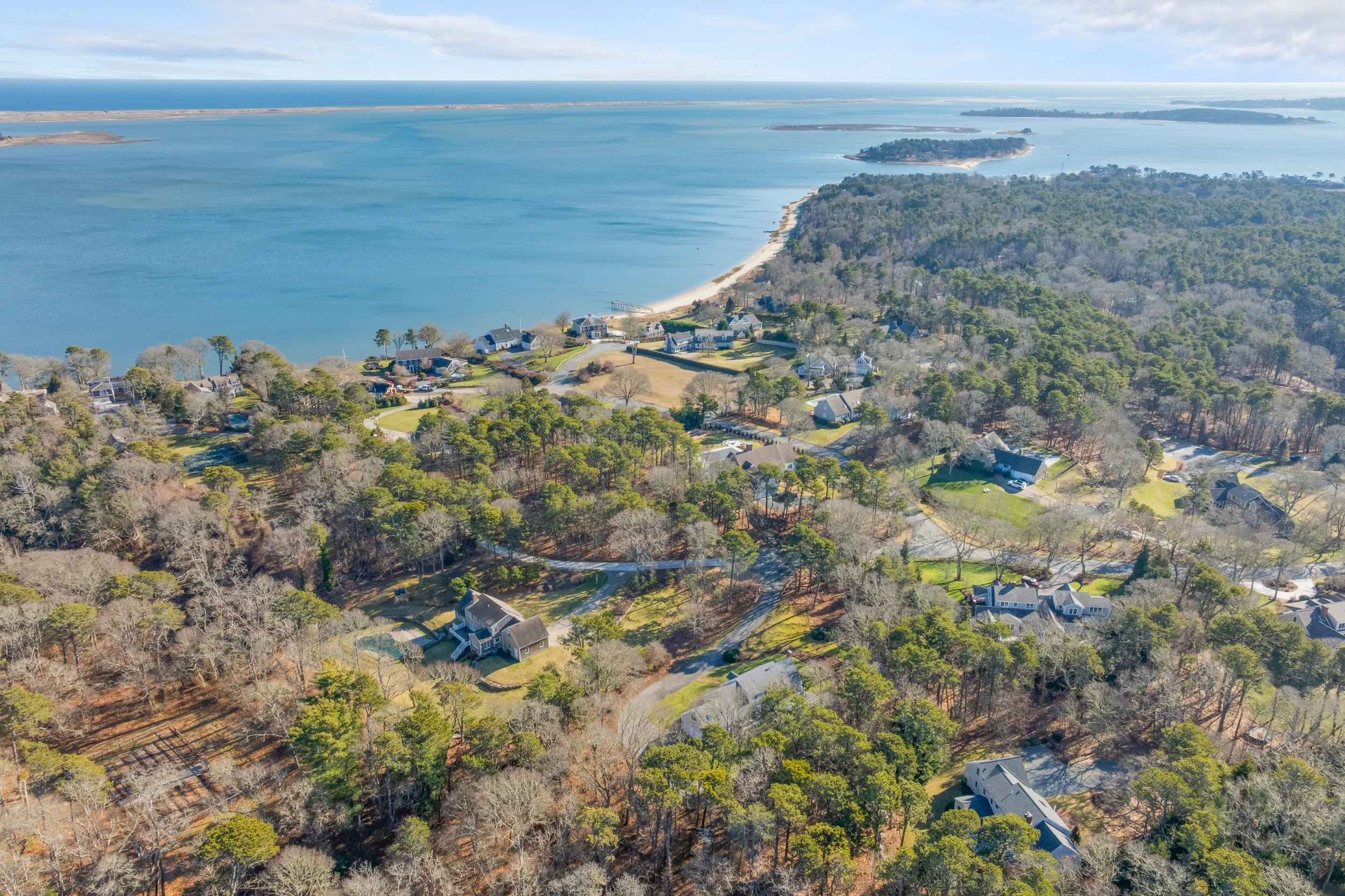 An aerial view of a coastal residential area with houses among trees, leading to a beach and body of water with small islands in the distance.