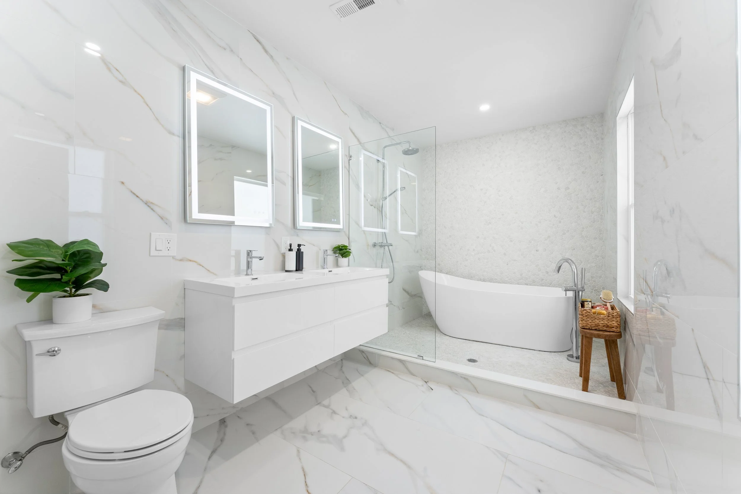 Modern bathroom with white marble walls and flooring, double vanity with mirrors, a bathtub, and a separate shower area with glass divider. Decor includes green plants and bathroom accessories.