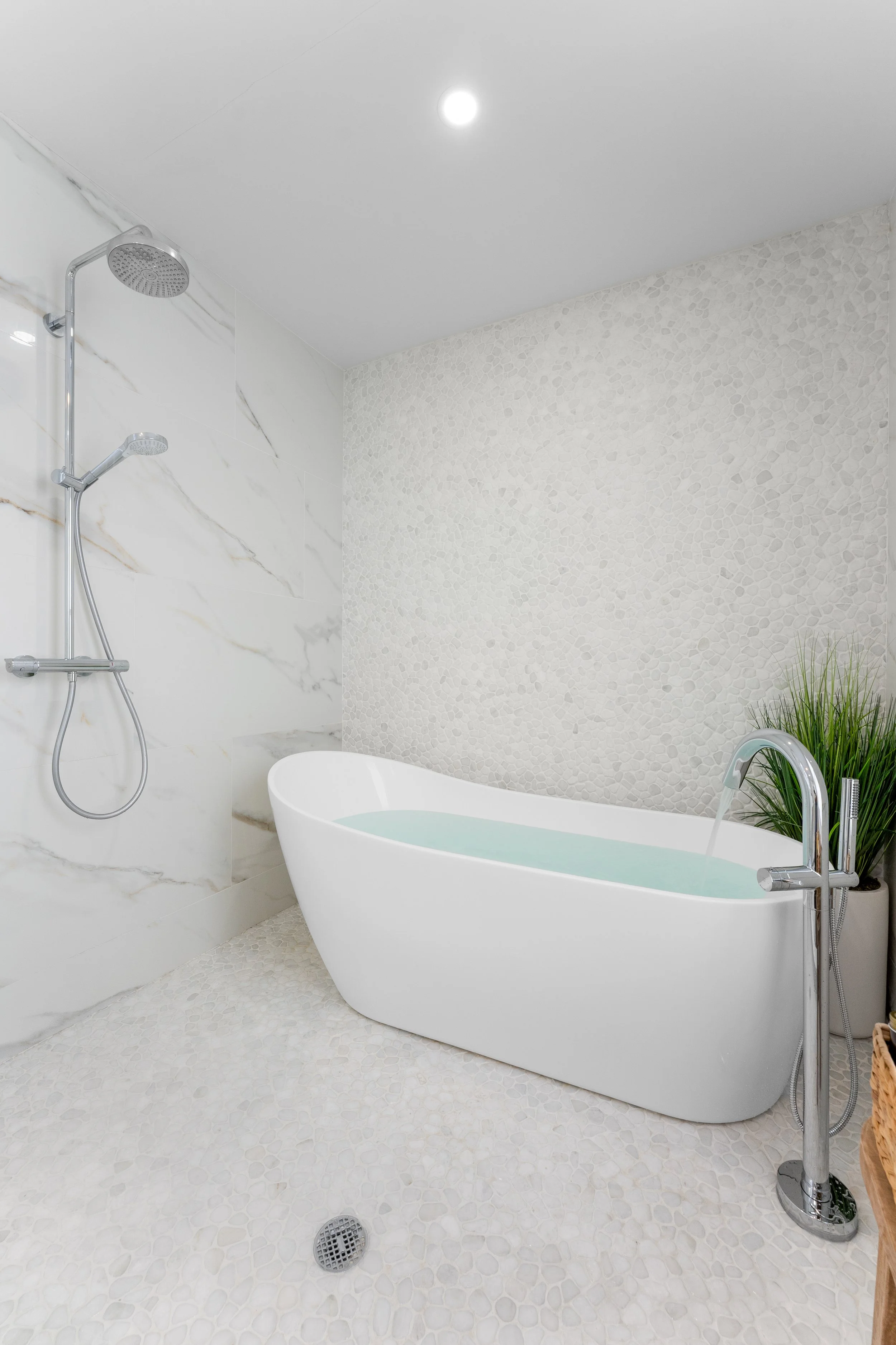 Modern bathroom featuring a white freestanding bathtub, a wall-mounted shower with a handheld showerhead, marble and pebble tile walls, and a small plant in a white container