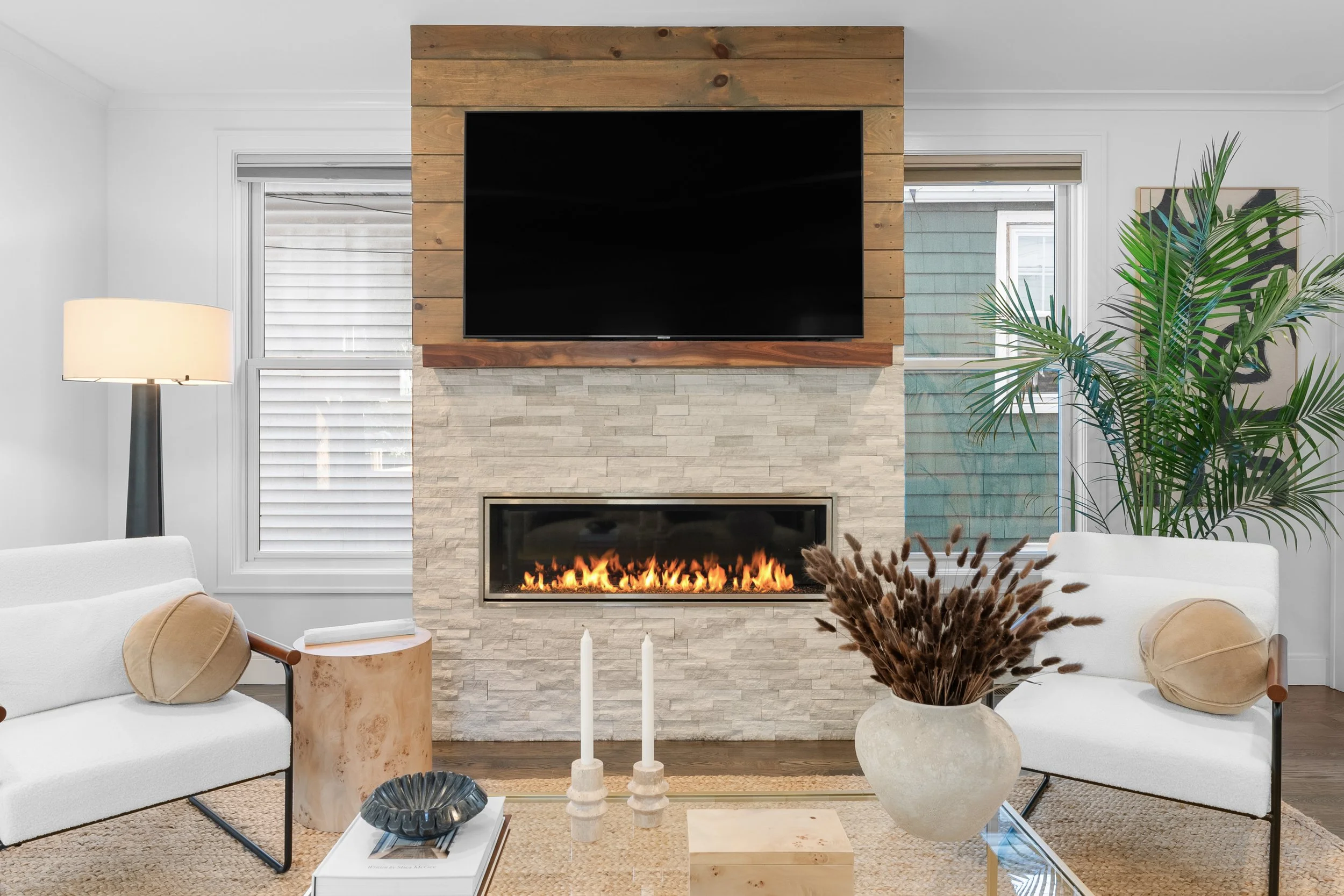 Living room with a fireplace and a large flat-screen TV mounted above, surrounded by a wood-paneled accent wall, with two white sofas, a beige round side table, a tall floor lamp, plants, candles, and decorative objects.