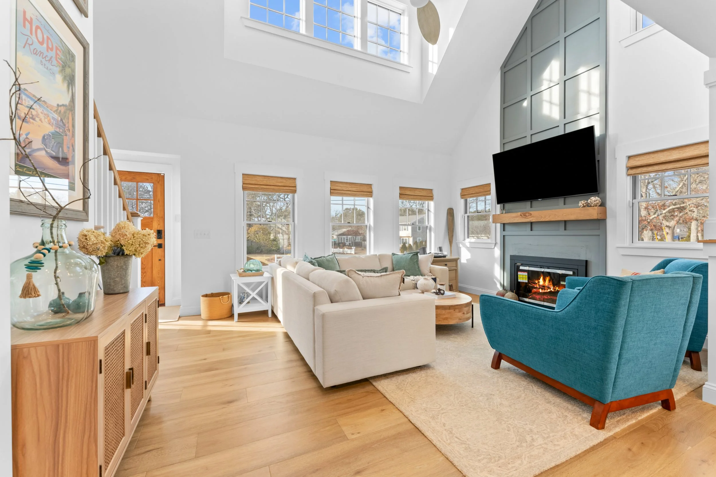 Bright living room with large windows, a fireplace, a beige sofa, a teal armchair, and decorative items on a wooden sideboard.