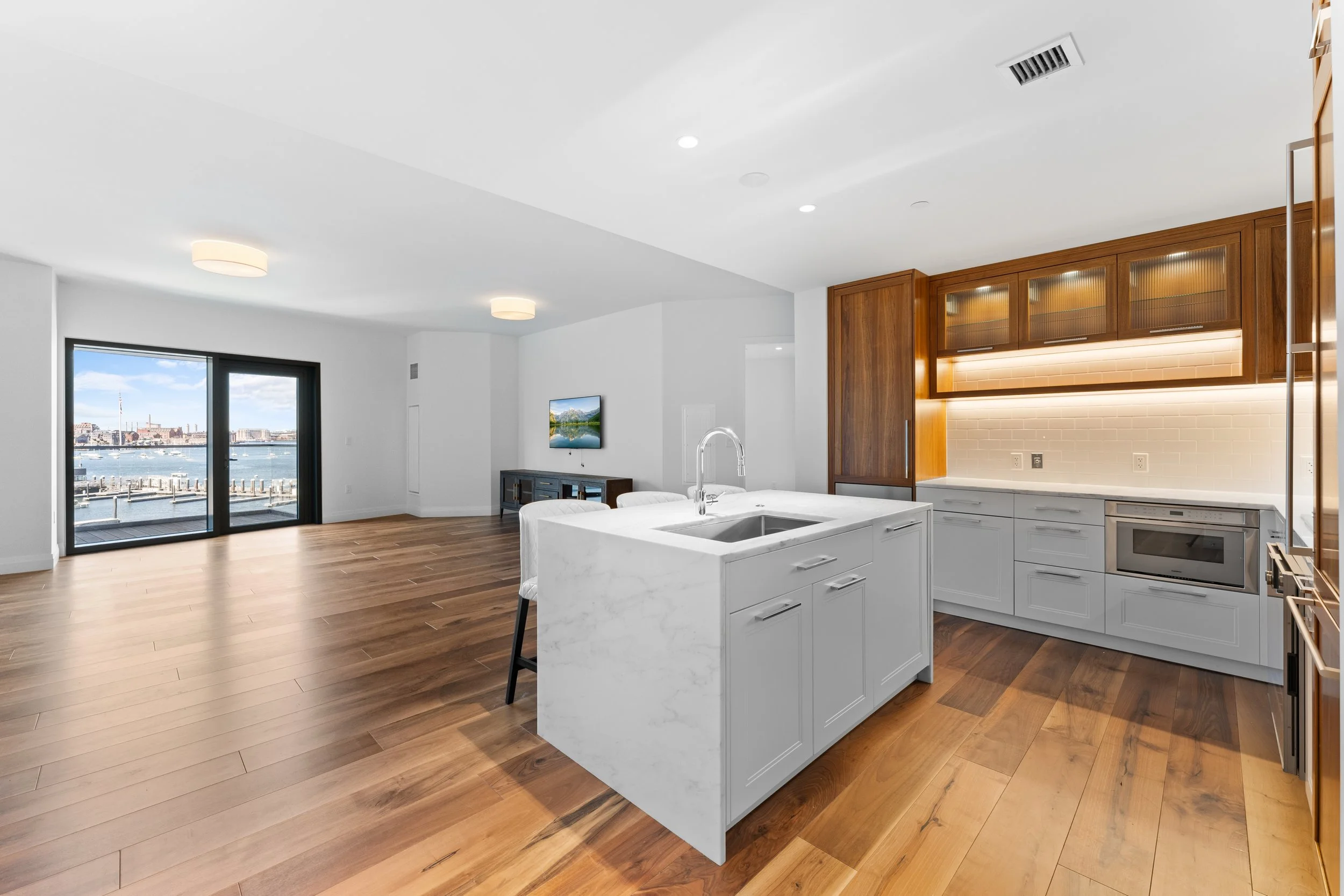 Open concept living space with hardwood floors, white walls, kitchen with island, wooden cabinets, and a view of a waterfront through glass doors.