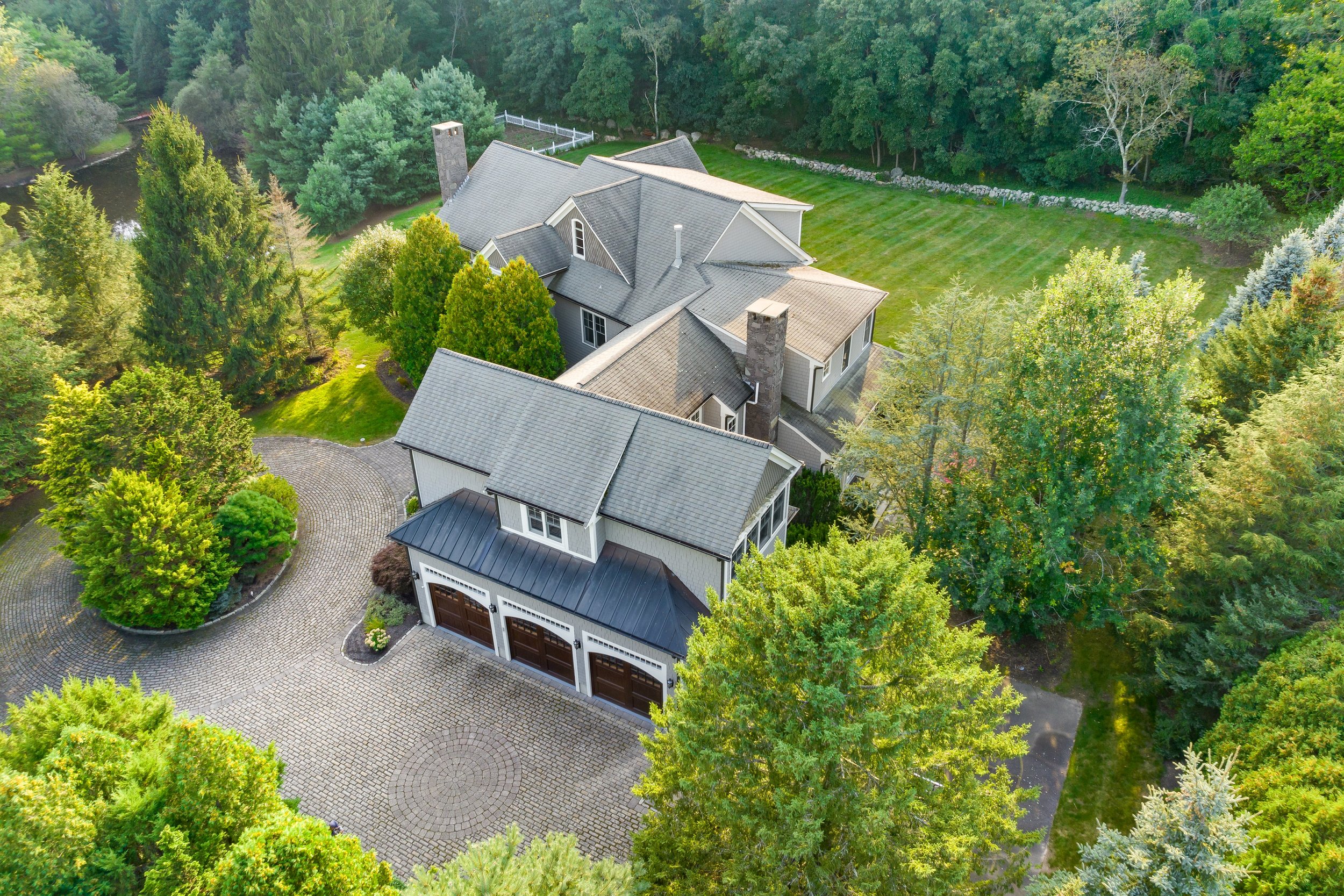 Aerial view of a large house with a gray roof and white exterior, surrounded by a lush green landscape with tall trees, a manicured lawn, and a paved driveway with a circular design.