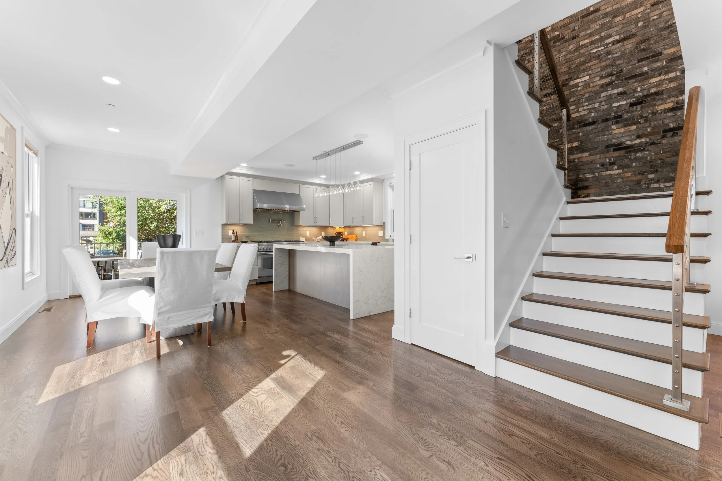 Bright, open-concept kitchen and dining area with white walls, wooden flooring, a staircase with dark wood steps and a brick accent wall, large windows, and minimalist furnishings.