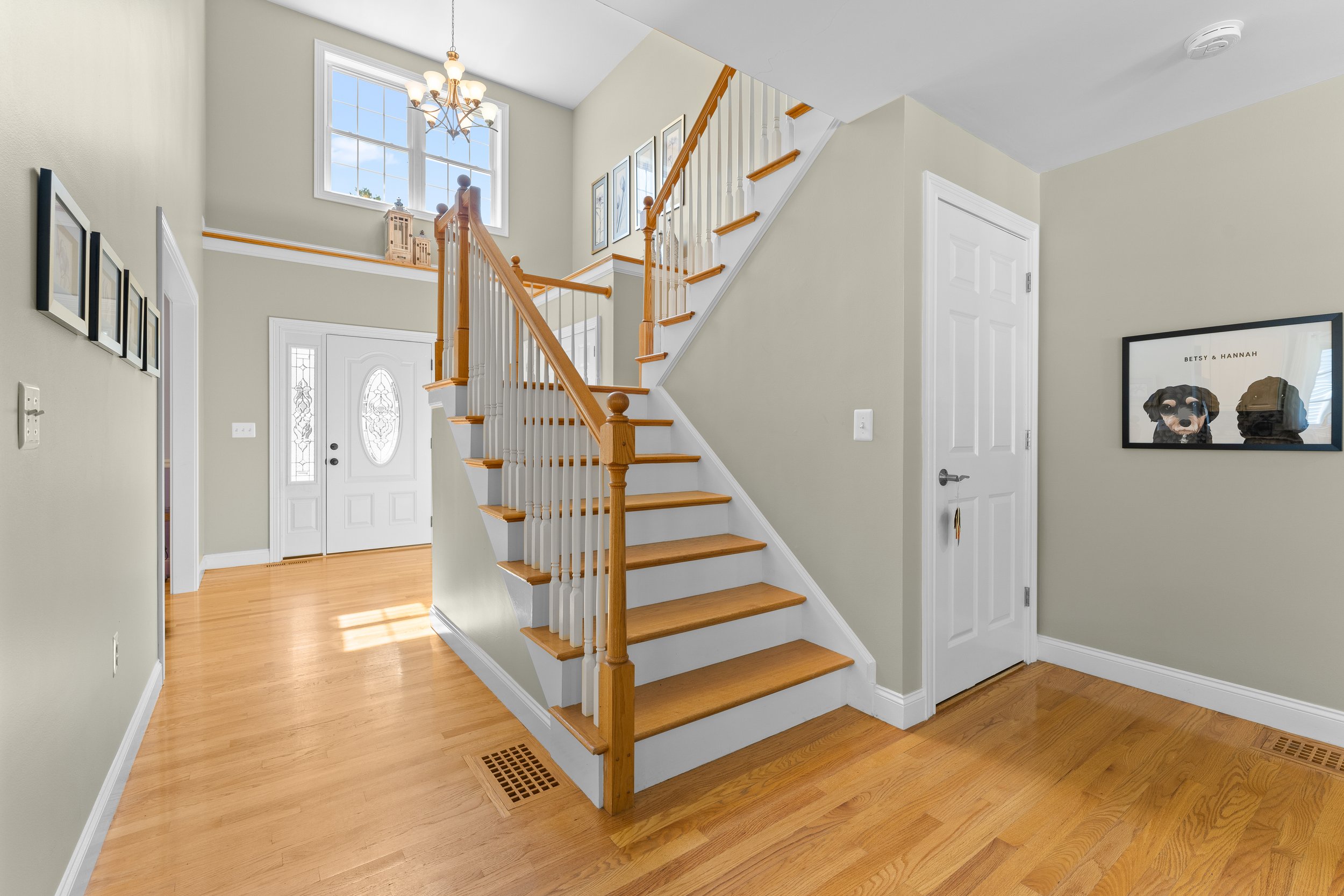 Interior view of a house entryway with hardwood flooring, a staircase with wooden handrails and white balusters, a front door with decorative glass, large windows overhead, and framed wall art.