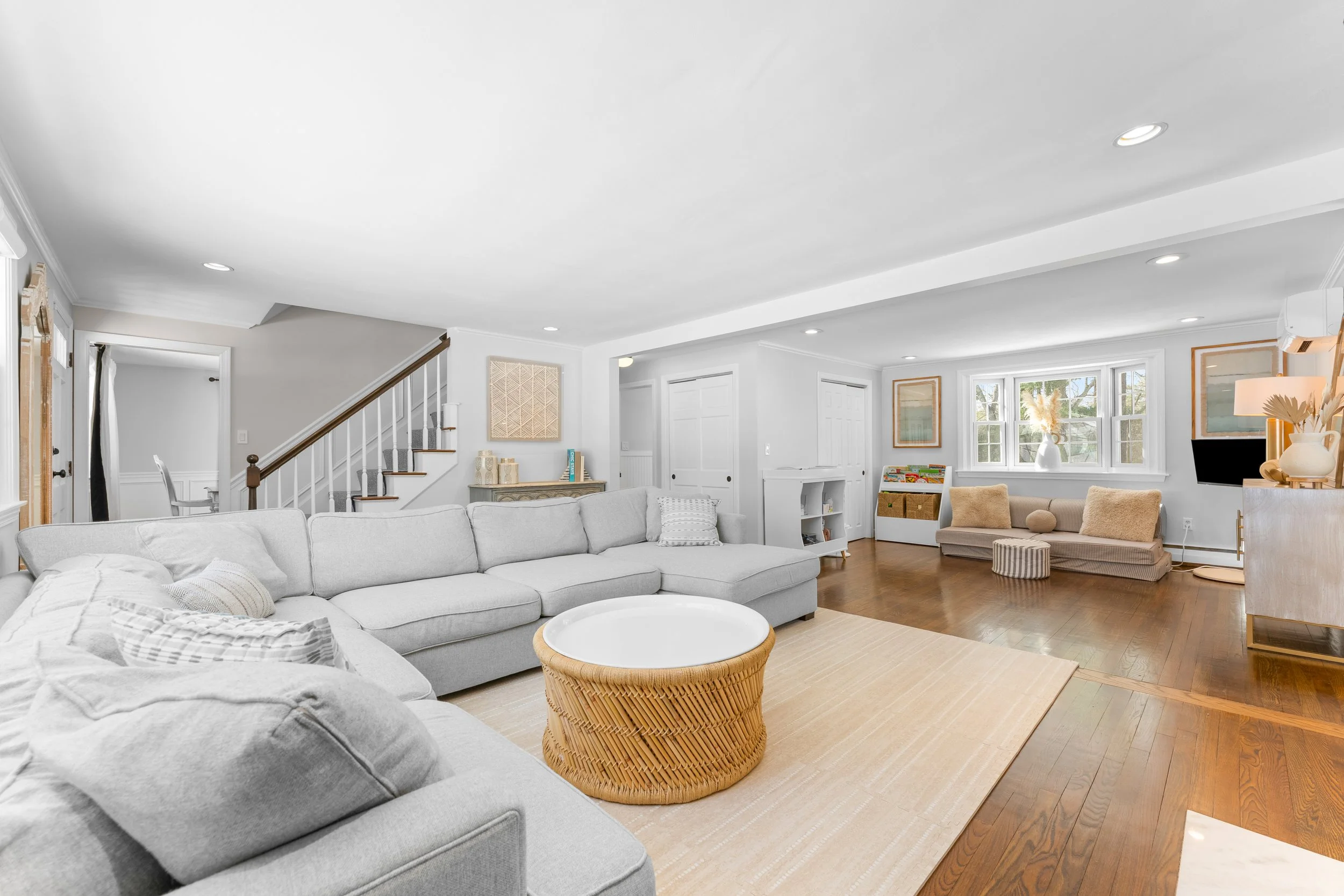 Bright spacious living room with large white sectional sofa, beige and striped pillows, a wicker coffee table, light hardwood floors, and decor including artwork, vases, and a television.