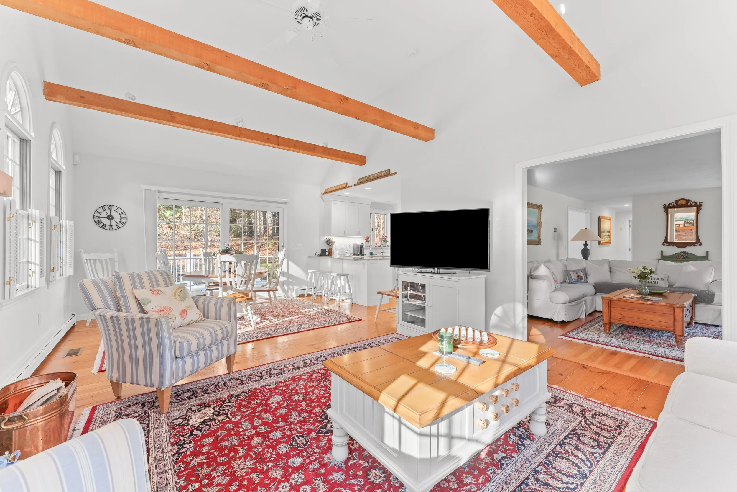 Interior of a bright living room with white walls, wooden beams, and hardwood floors. Contains striped armchairs, a white sofa, a wooden coffee table, a large area rug, and a mounted flat-screen TV. Large windows show a wooded outdoor scene.