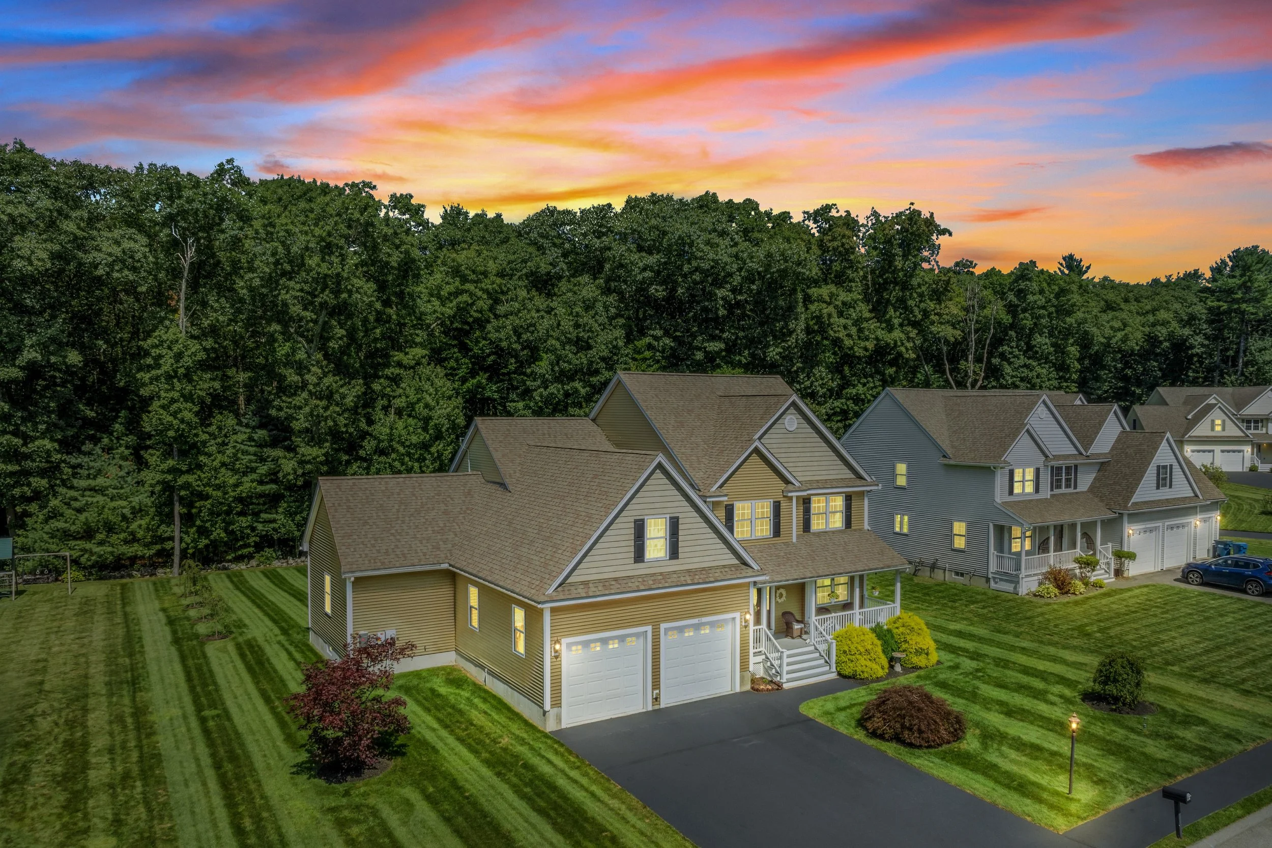 A neighborhood scene at dusk with multiple houses, manicured lawns, and trees, under a colorful sky with pink, orange, and blue clouds.