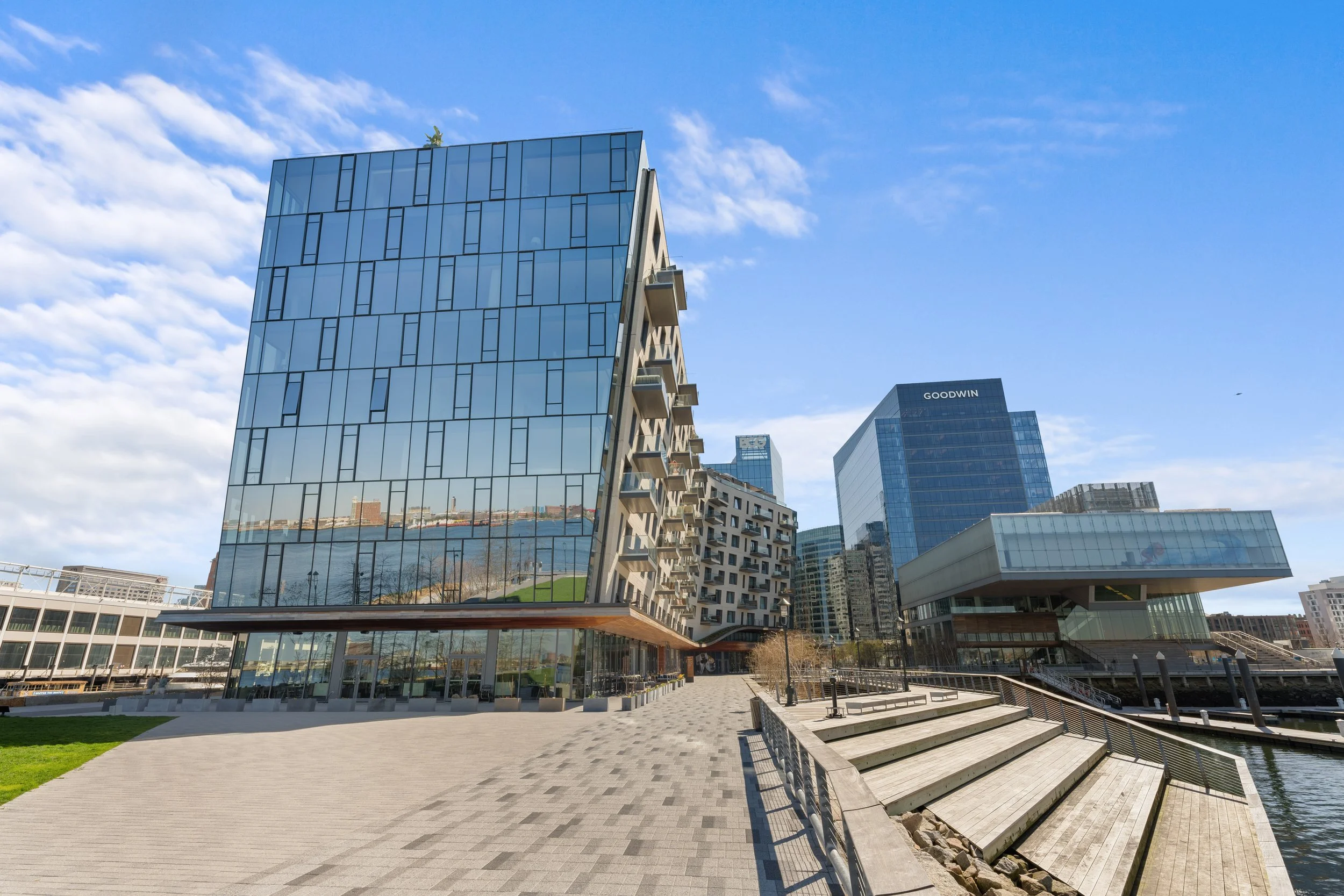 Modern glass office buildings with a waterfront walkway, steps, and a bridge, under a partly cloudy sky.
