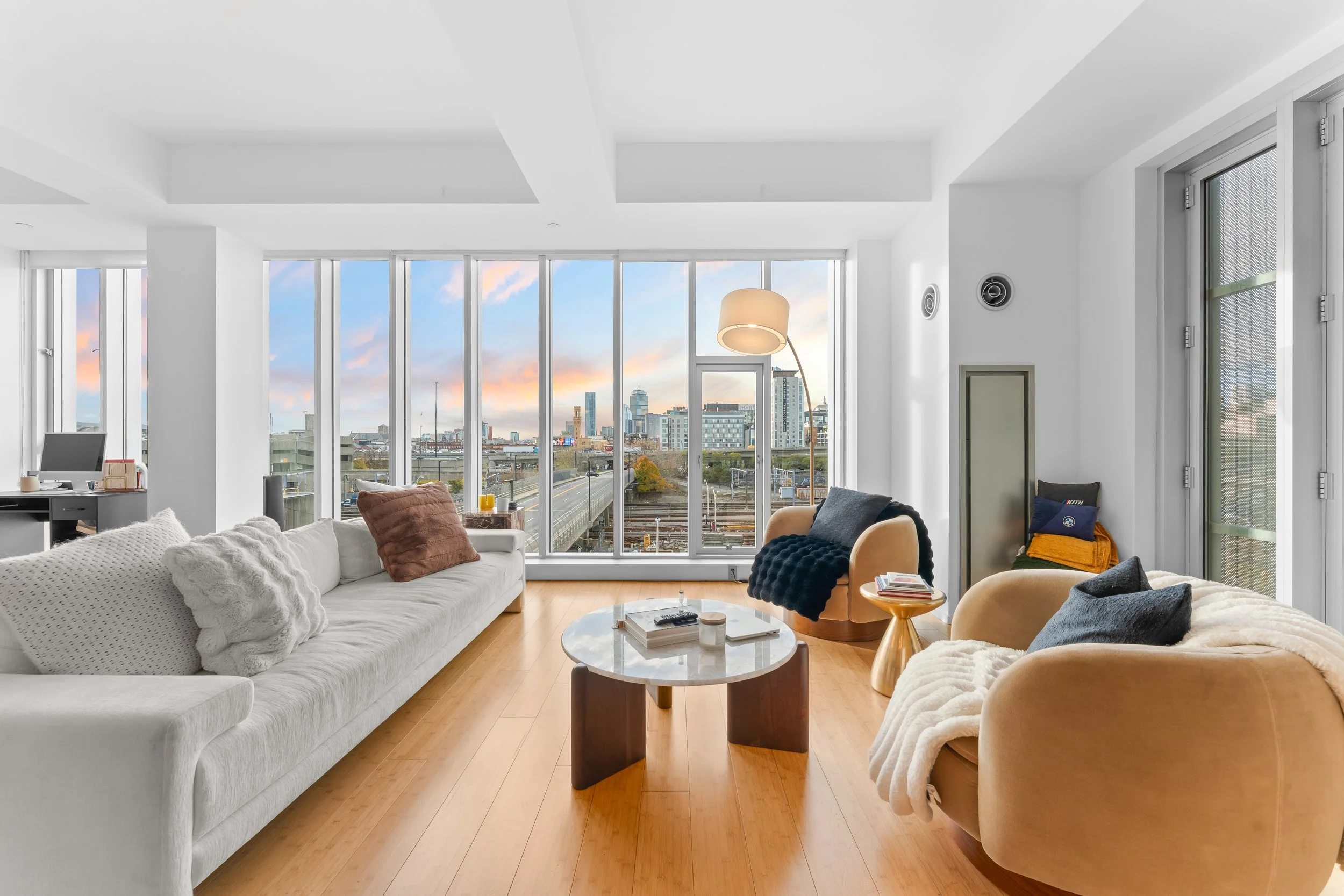 Bright modern living room with large windows showing cityscape and sky, white sofa with throw pillows, beige armchairs, glass coffee table, and wooden flooring.