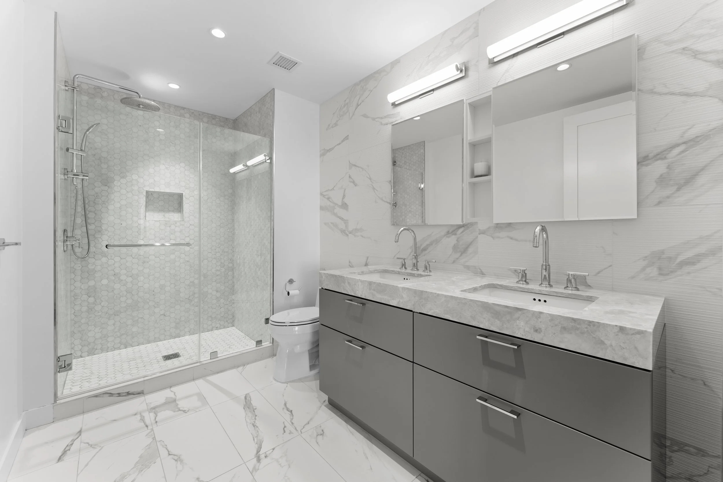 Modern bathroom with a glass-enclosed shower, a white toilet, marble tiles on the walls and floor, and a double sink vanity with gray cabinets and illuminated mirrors.