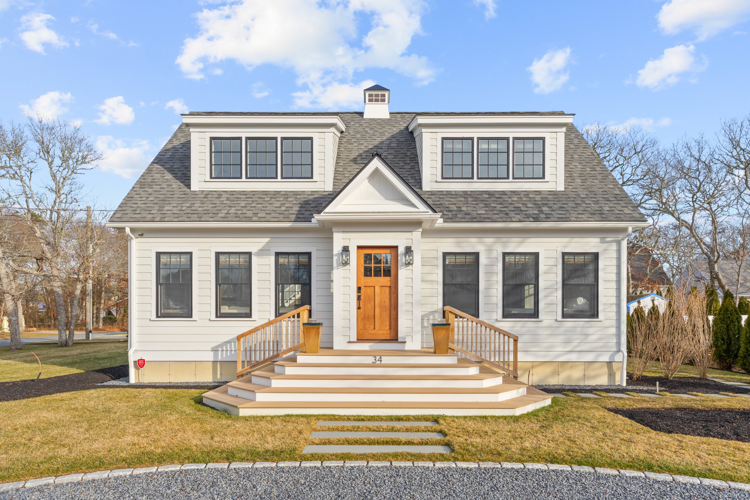 Front view of a white, two-story house with a gabled roof, dormer windows, and a wooden front door with steps leading up to it, set against a partly cloudy sky with leafless trees nearby.
