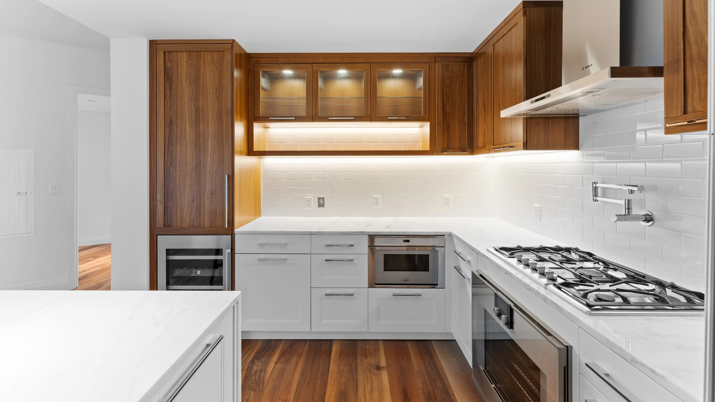 A modern kitchen with white lower cabinets, gray marble countertops, and wooden upper cabinets with glass doors. Stainless steel appliances include a gas stove, oven, and wine cooler. The backsplash features white subway tiles, and there is under-cabinet lighting.