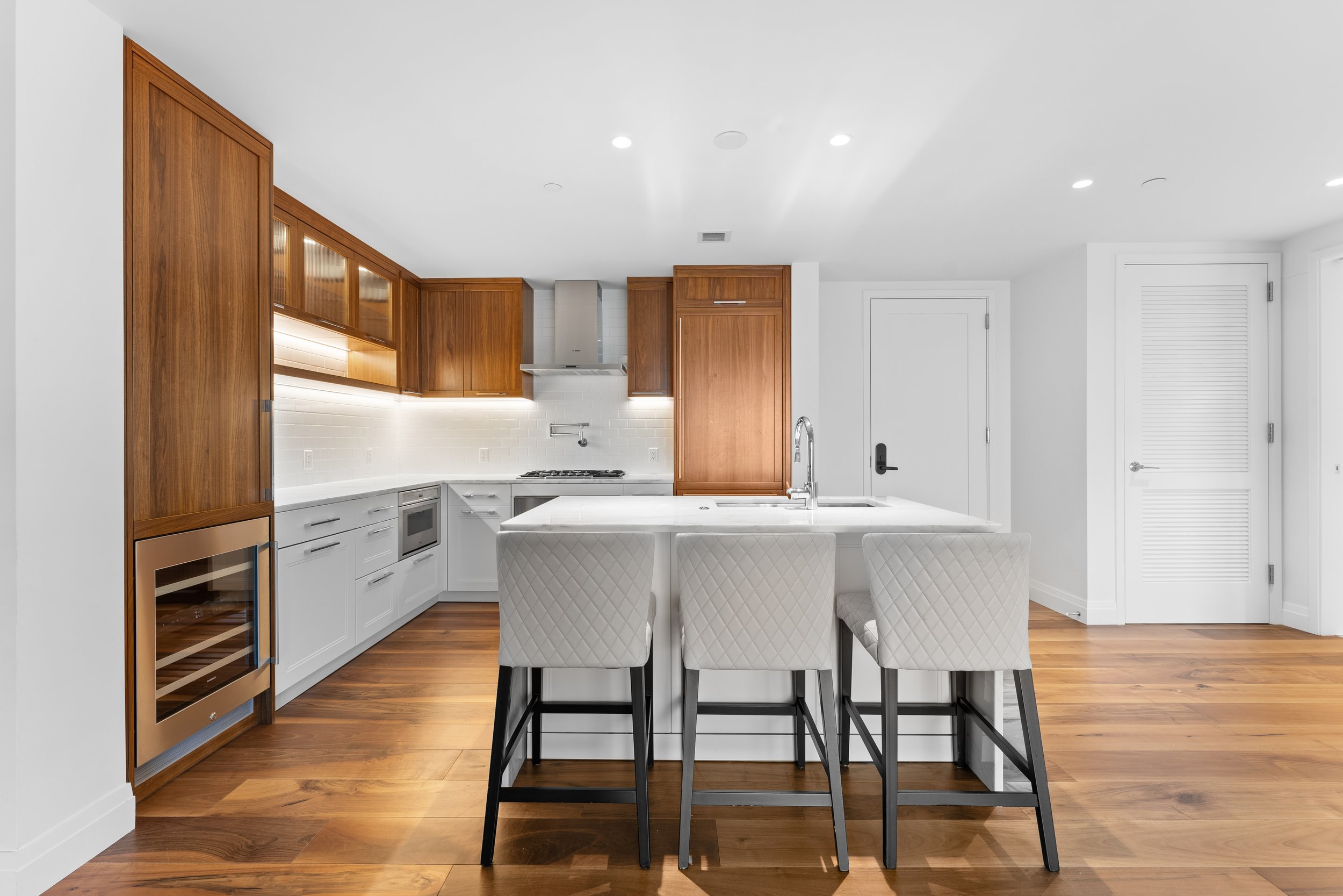 Modern kitchen with white cabinets, wood accents, and a white marble island with quilted white dining chairs.