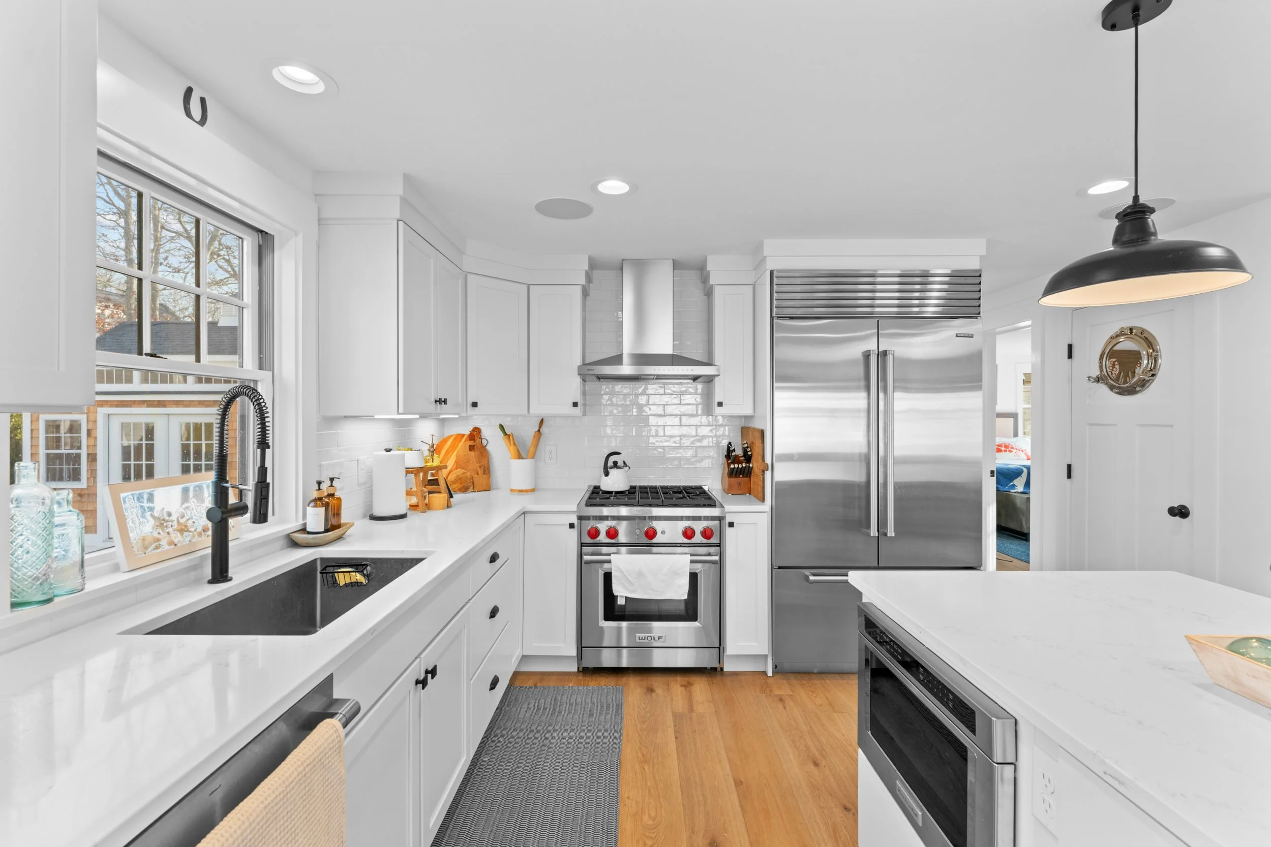 A modern bright kitchen with white cabinets, a large window above the sink, stainless steel appliances including a refrigerator and a stove, wooden countertops, and various kitchen accessories and utensils.