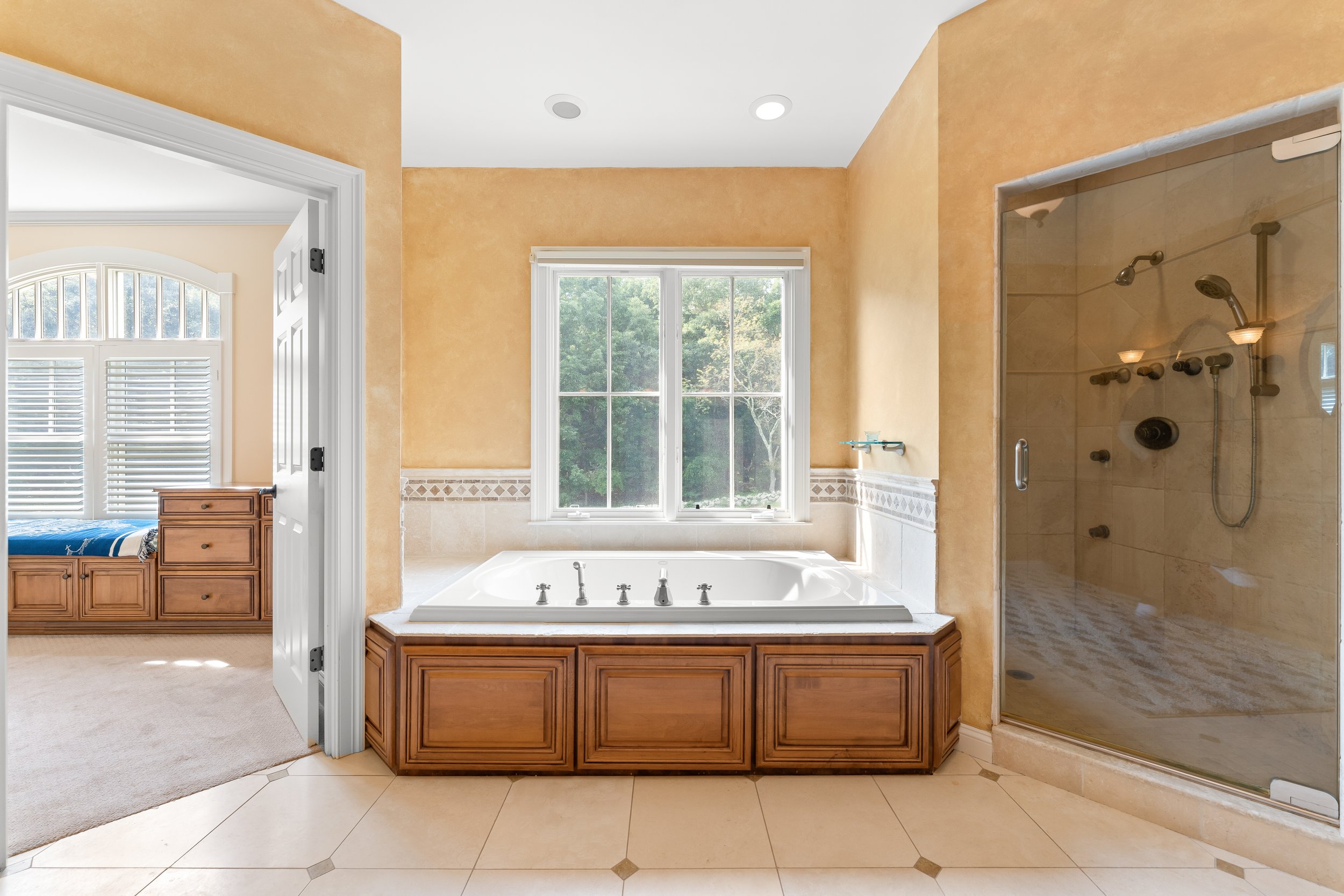 Bathroom with large window above a jacuzzi tub, a glass-enclosed shower, and a bedroom door visible on the left.