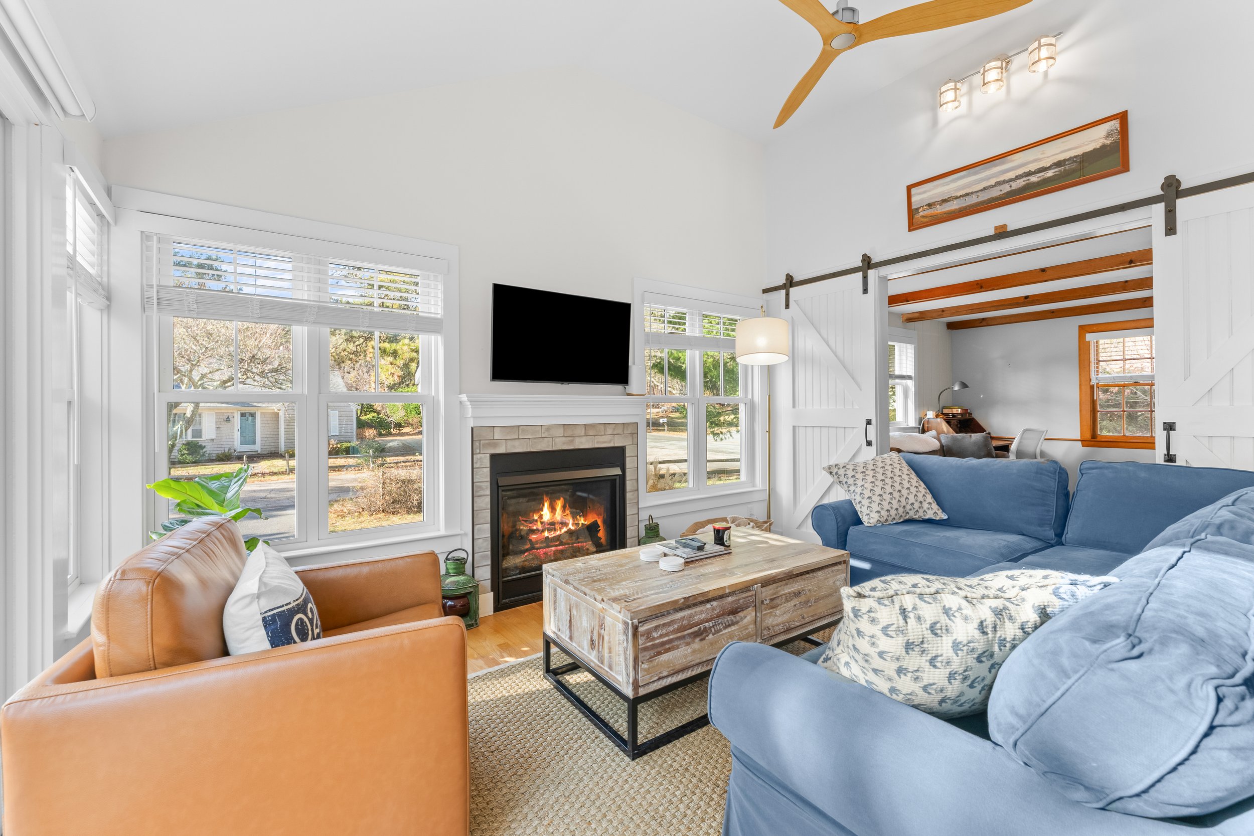 Living room with large windows, white walls, a fireplace, flat-screen TV, wooden ceiling beams, a wood coffee table, and a mix of blue and beige sofas and chairs.
