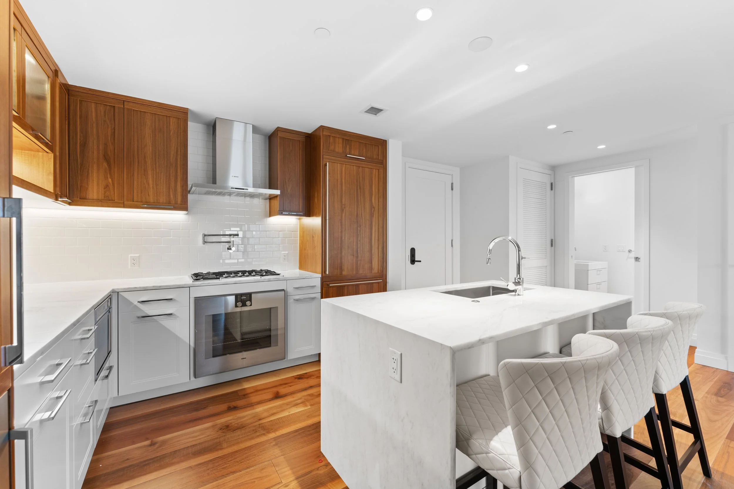 Modern kitchen with white cabinetry, wooden upper cabinets, white subway tile backsplash, stainless steel appliances, a white marble island with seating, hardwood flooring, and recessed lighting.