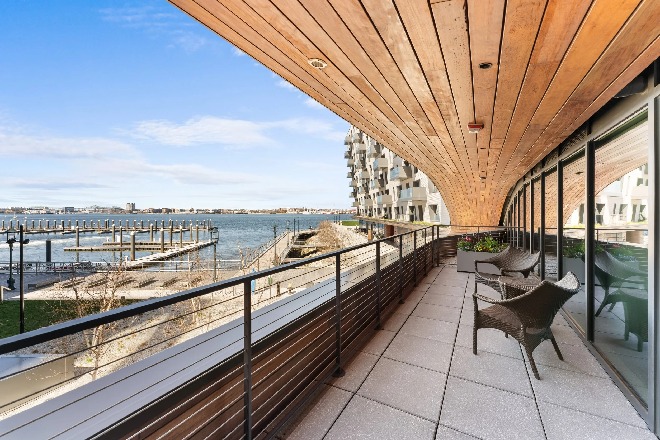 A balcony with a wooden ceiling and black chairs overlooking a body of water with boats and a city skyline in the distance.