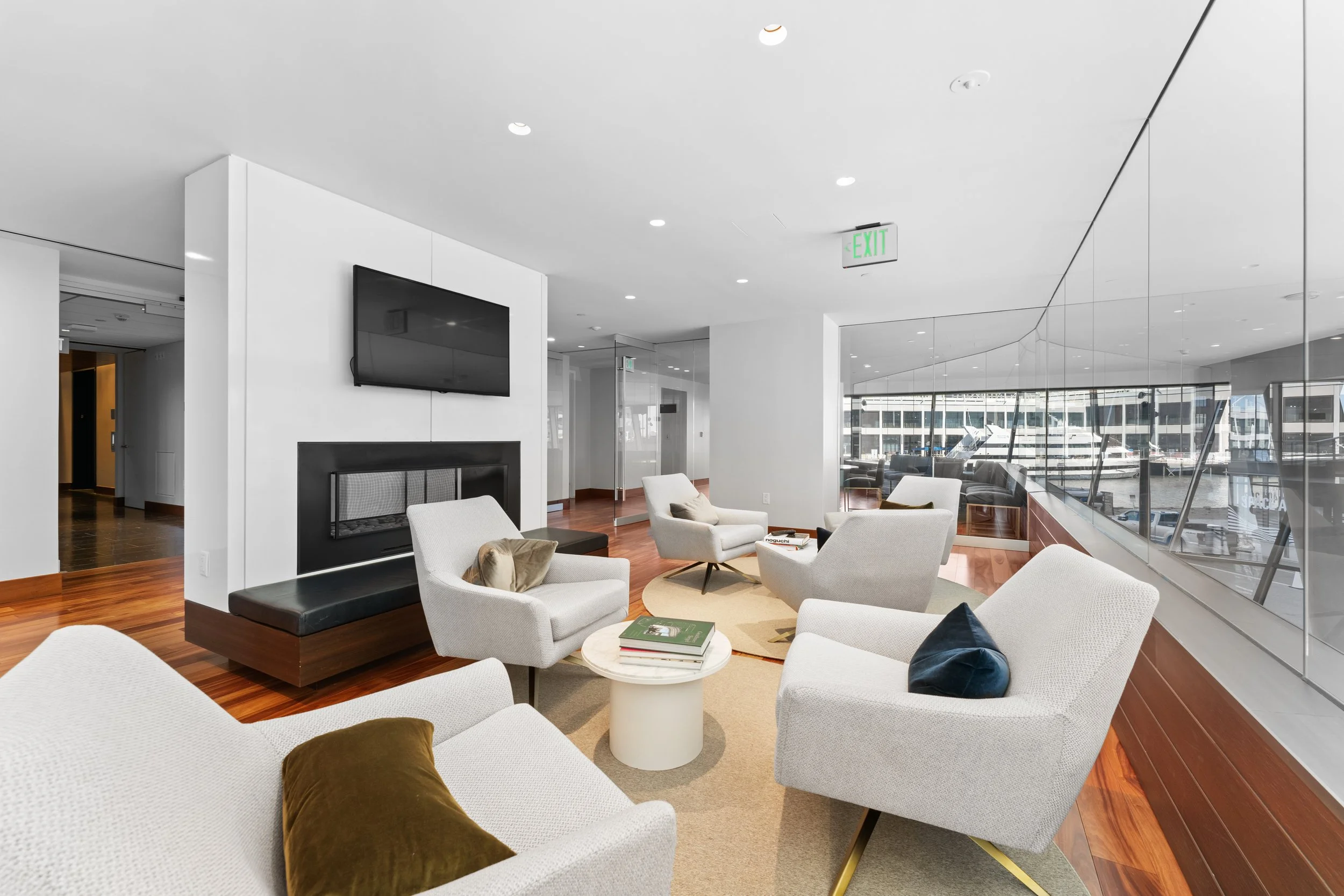 Modern lounge area with white armchairs, a beige rug, a small round table with magazines, a dark bench, a fireplace, a wall-mounted TV, large windows with a city and water view, and white walls and ceiling.