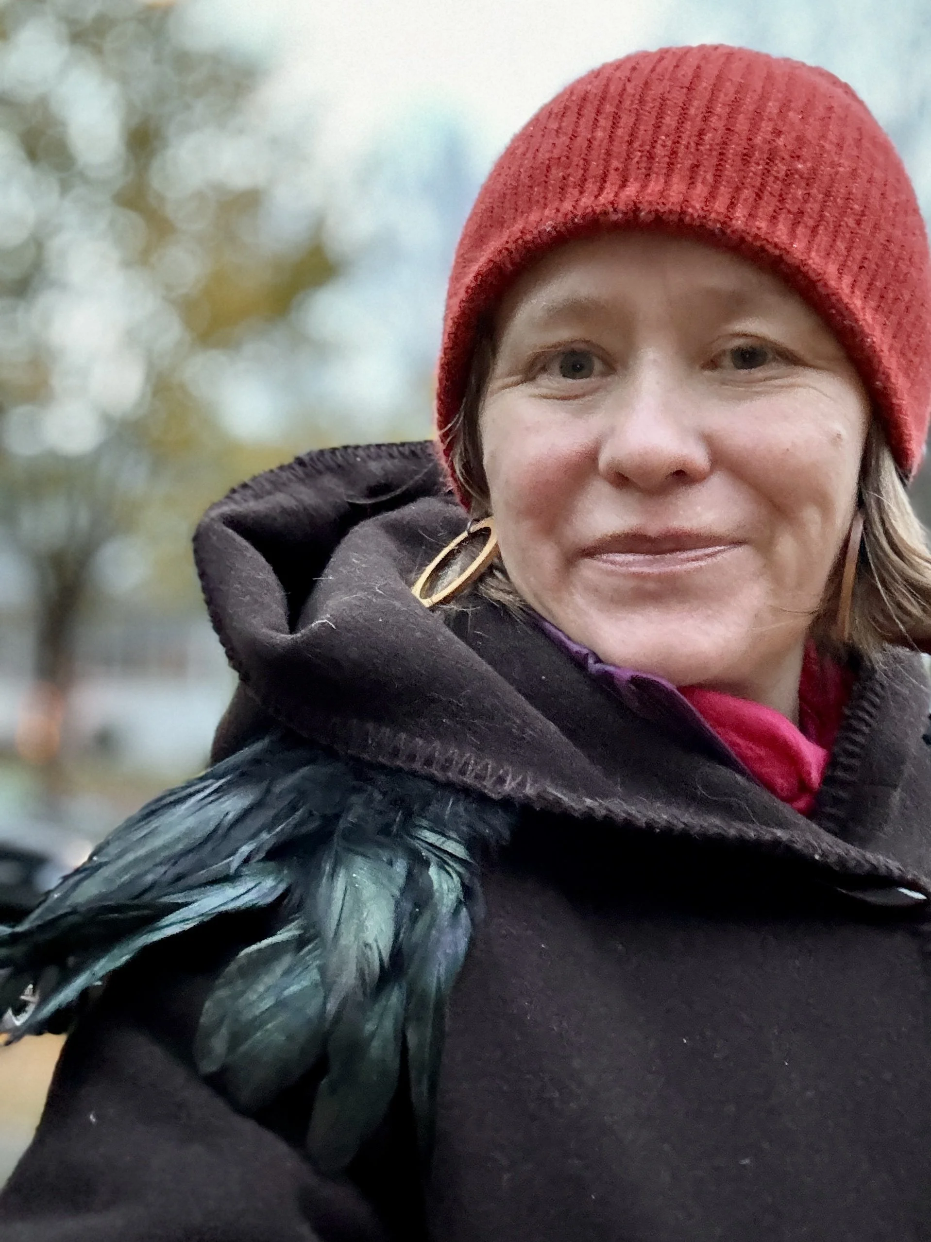 A woman wearing a red knit hat and black coat with blue-green feathers on the shoulder, standing outdoors with blurred trees in the background.