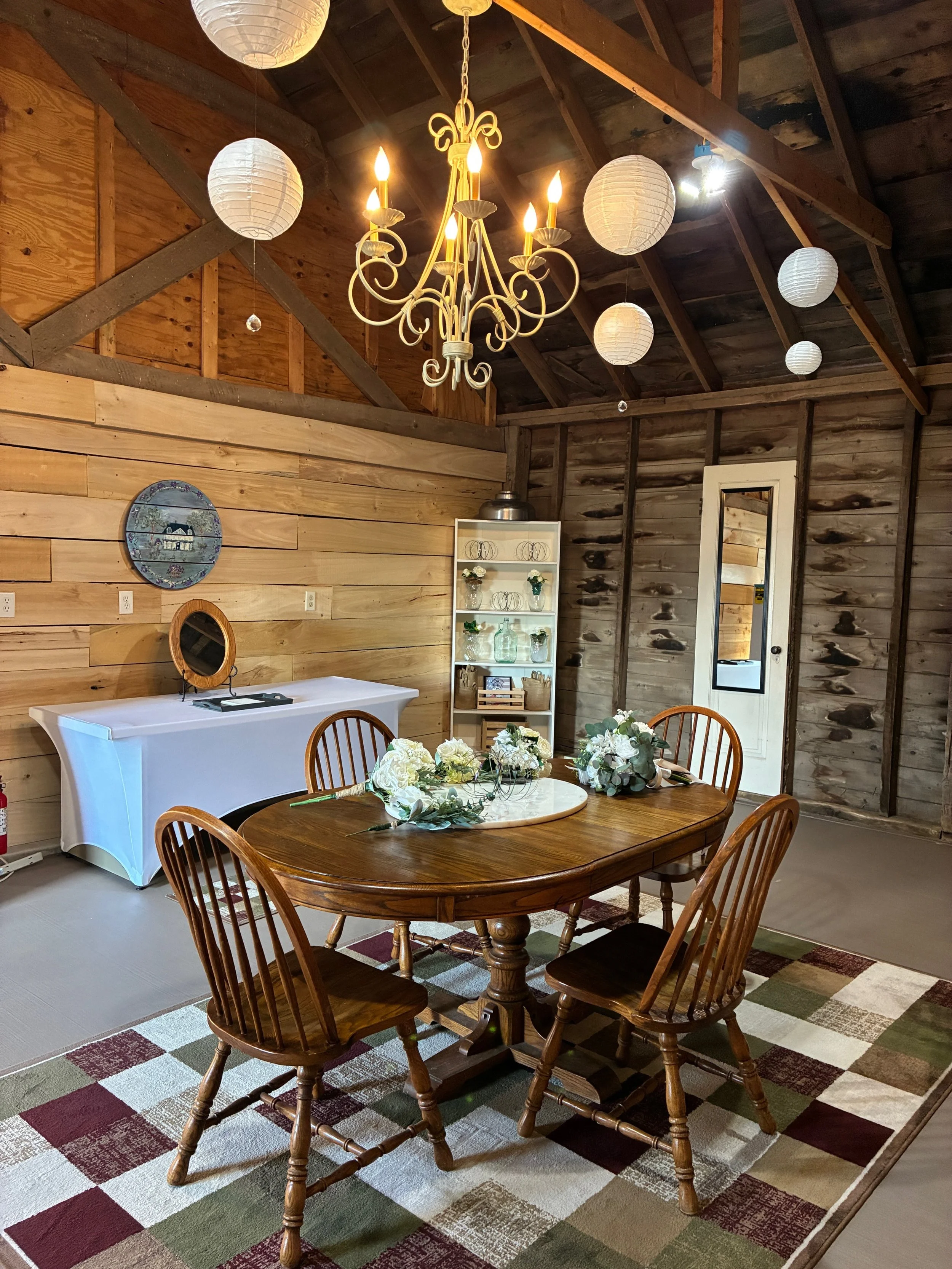 A rustic dining room with wooden walls and ceiling, a round wooden table with four chairs, floral centerpieces, a chandelier, paper lanterns, and a white sideboard with decor.