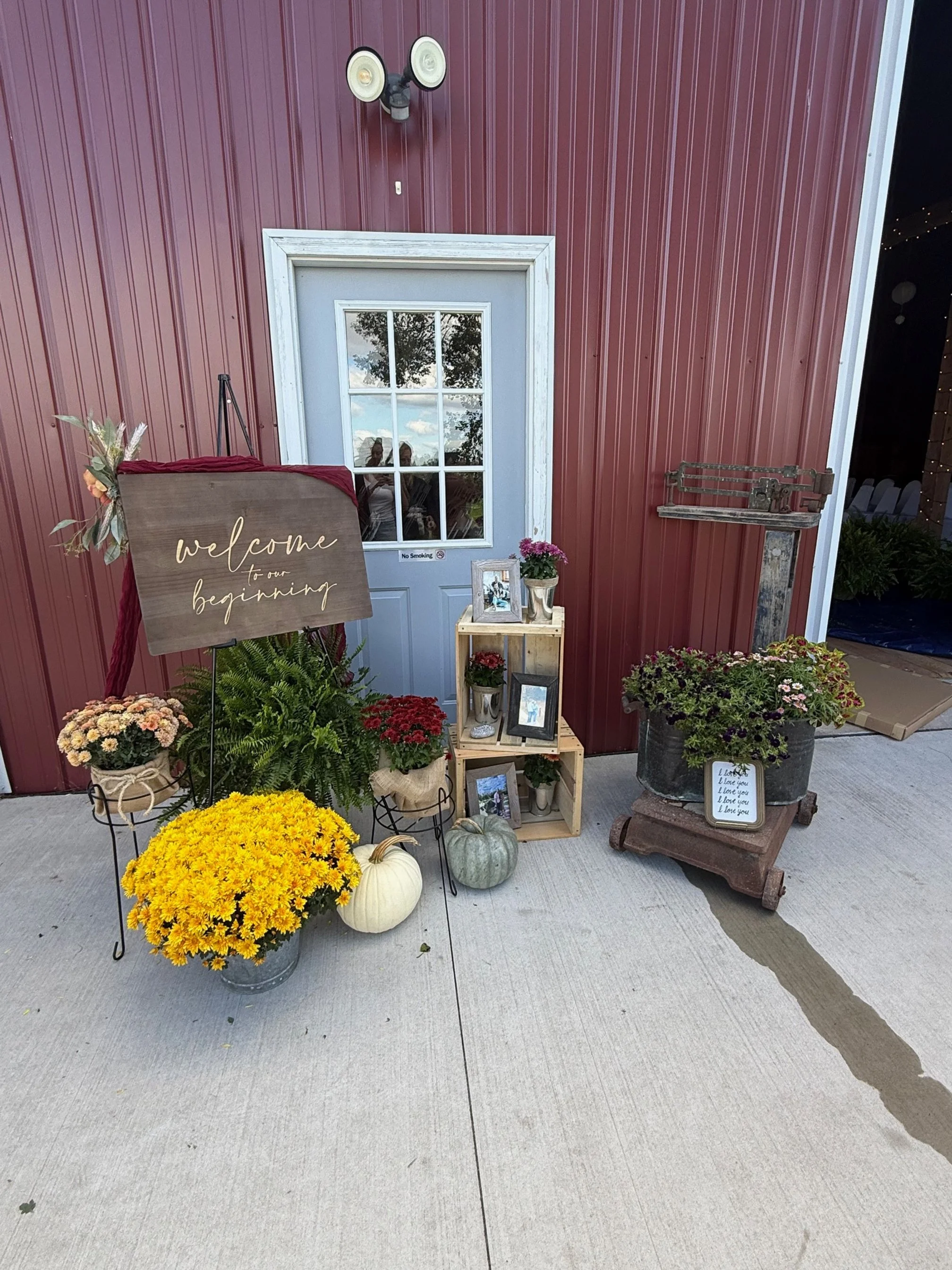 Outdoor floral display with a welcome sign, pumpkins, and potted plants arranged near a white door with windows, on a concrete surface.