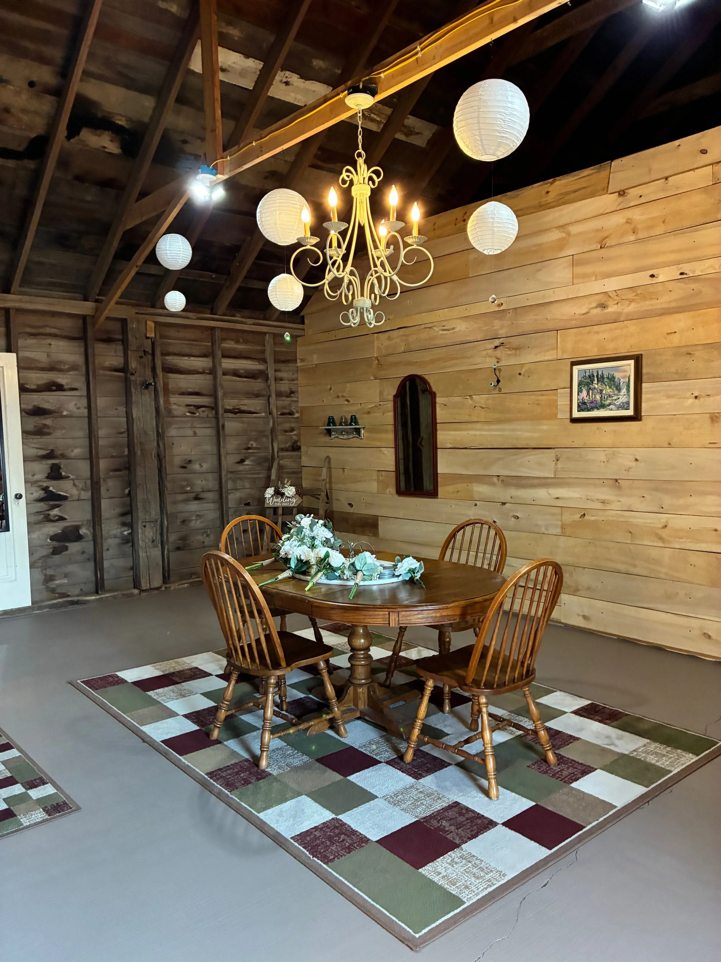 A rustic dining area with a wooden table and four matching chairs, a floral centerpiece, and a colorful checkered rug on a concrete floor. The room has wooden walls and ceiling, white paper lanterns hanging from the ceiling, and a vintage chandelier 