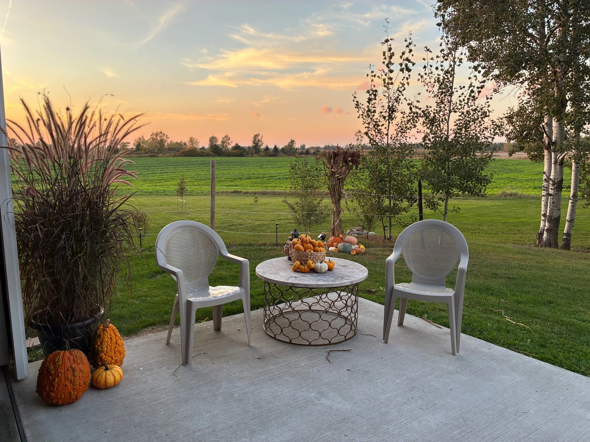 Patio with two white chairs and a round table decorated with pumpkins and gourds, overlooking a green lawn and trees at sunset.