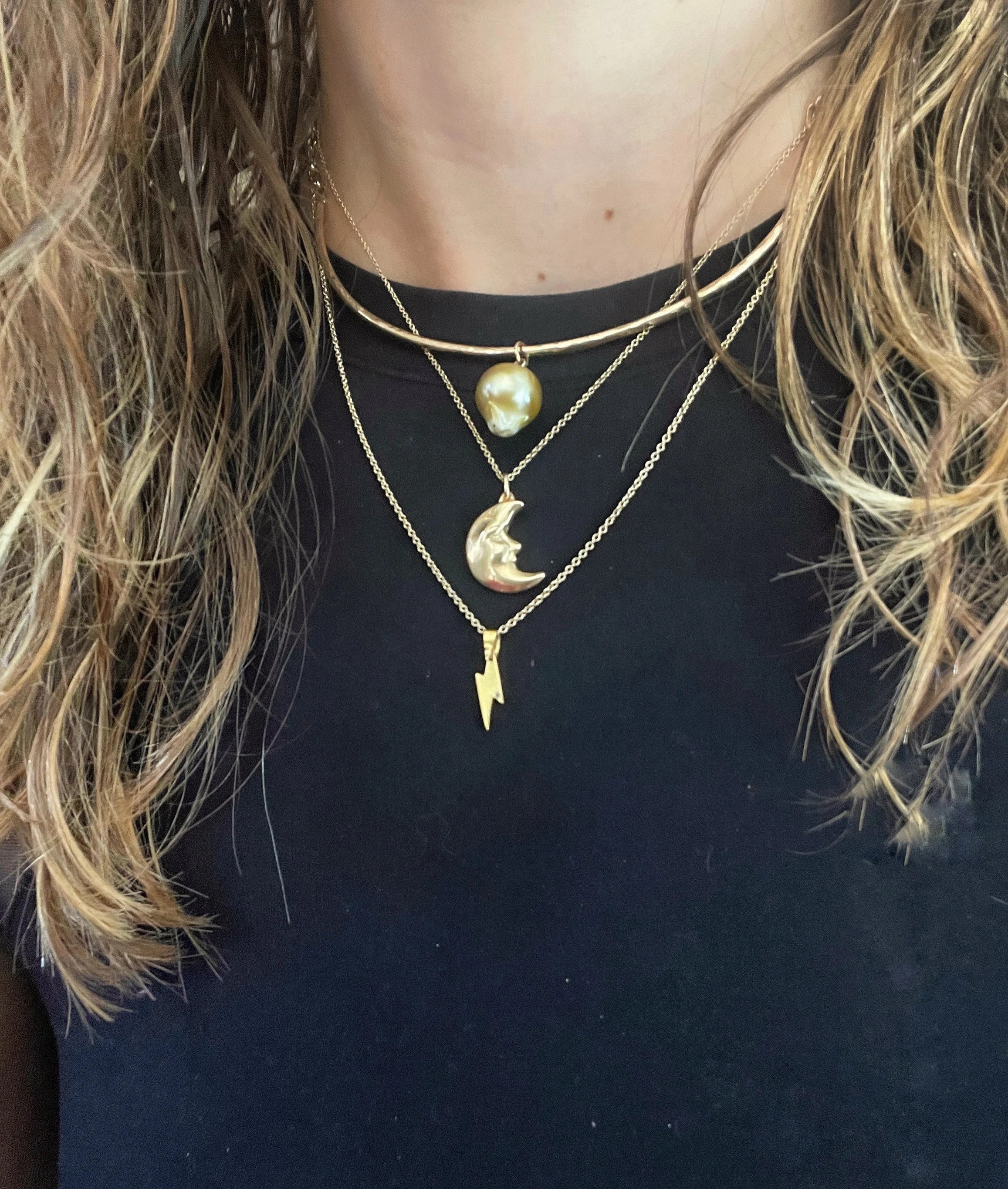Three layered gold necklaces with pearl, moon, and lightning bolt pendants worn over a black top.