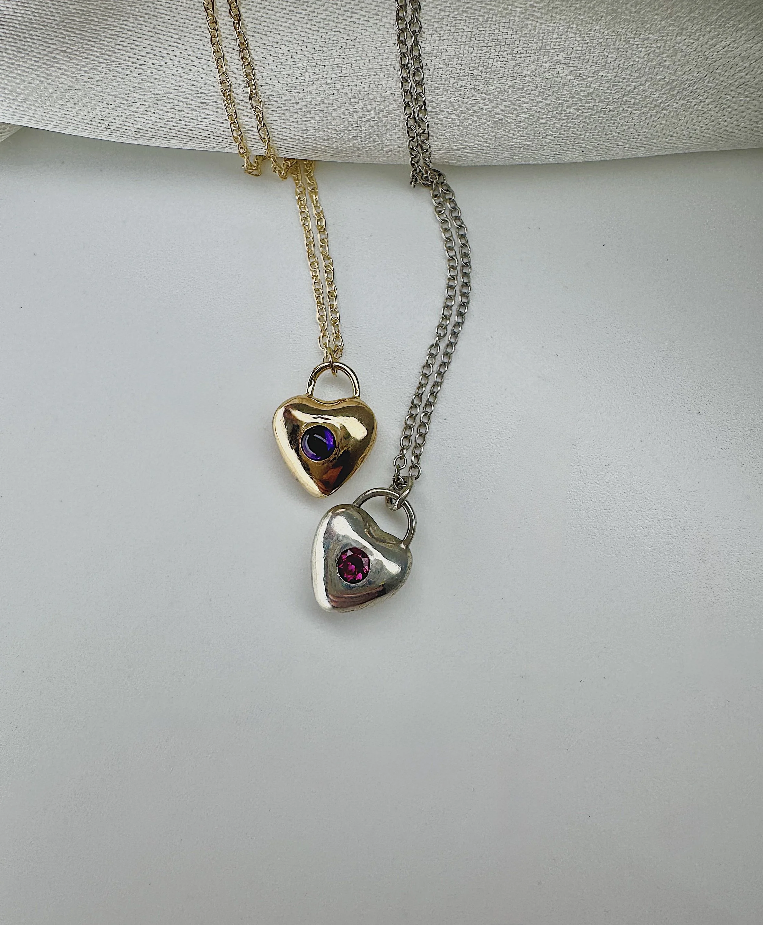 Two necklaces hanging from a cushion, one gold with a heart-shaped pendant and one silver with a similar heart pendant, both with purple gemstones.