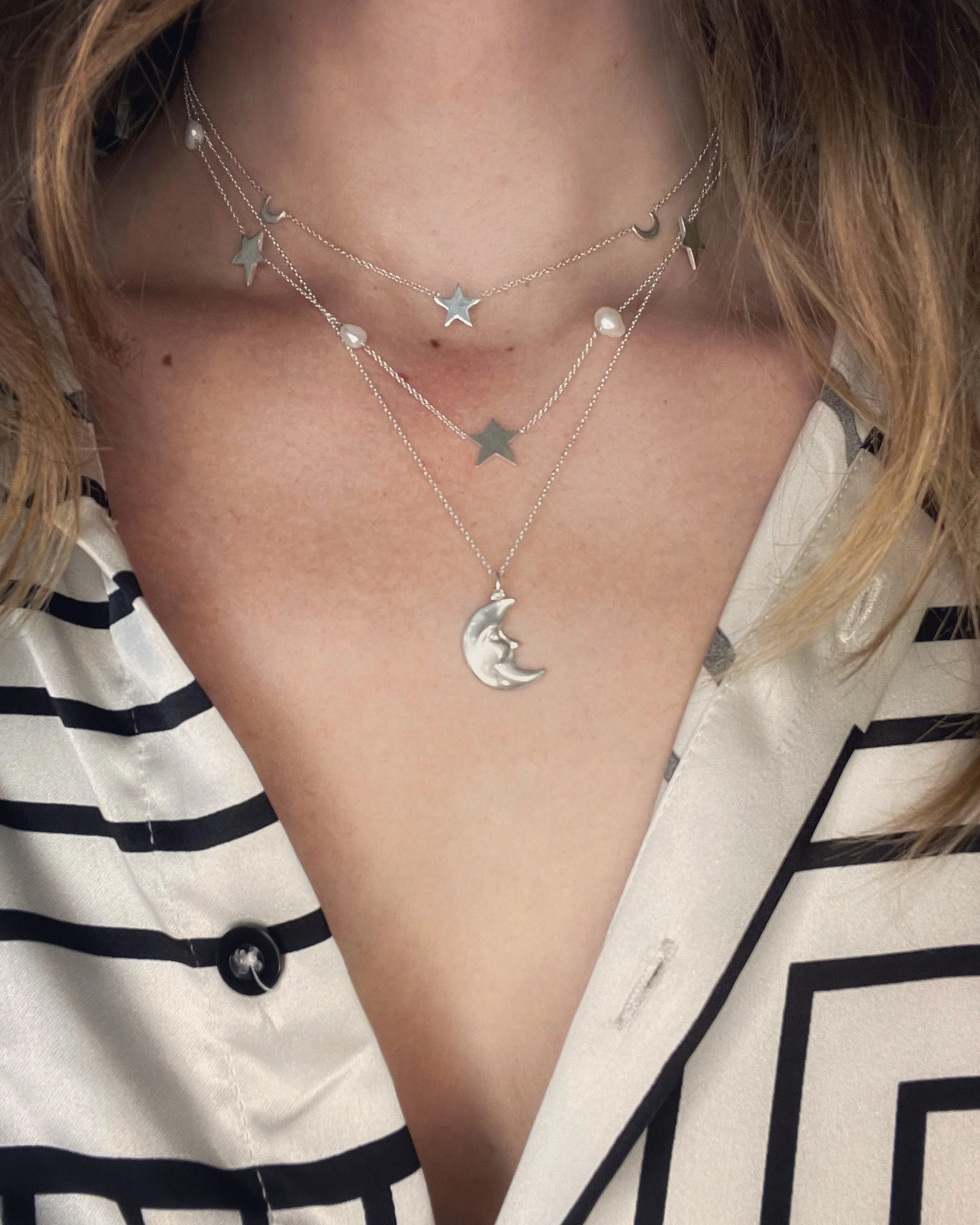 Close-up of a woman's neck and chest showing layered silver necklaces with star, moon, and pearl pendants, wearing a black and white striped shirt.
