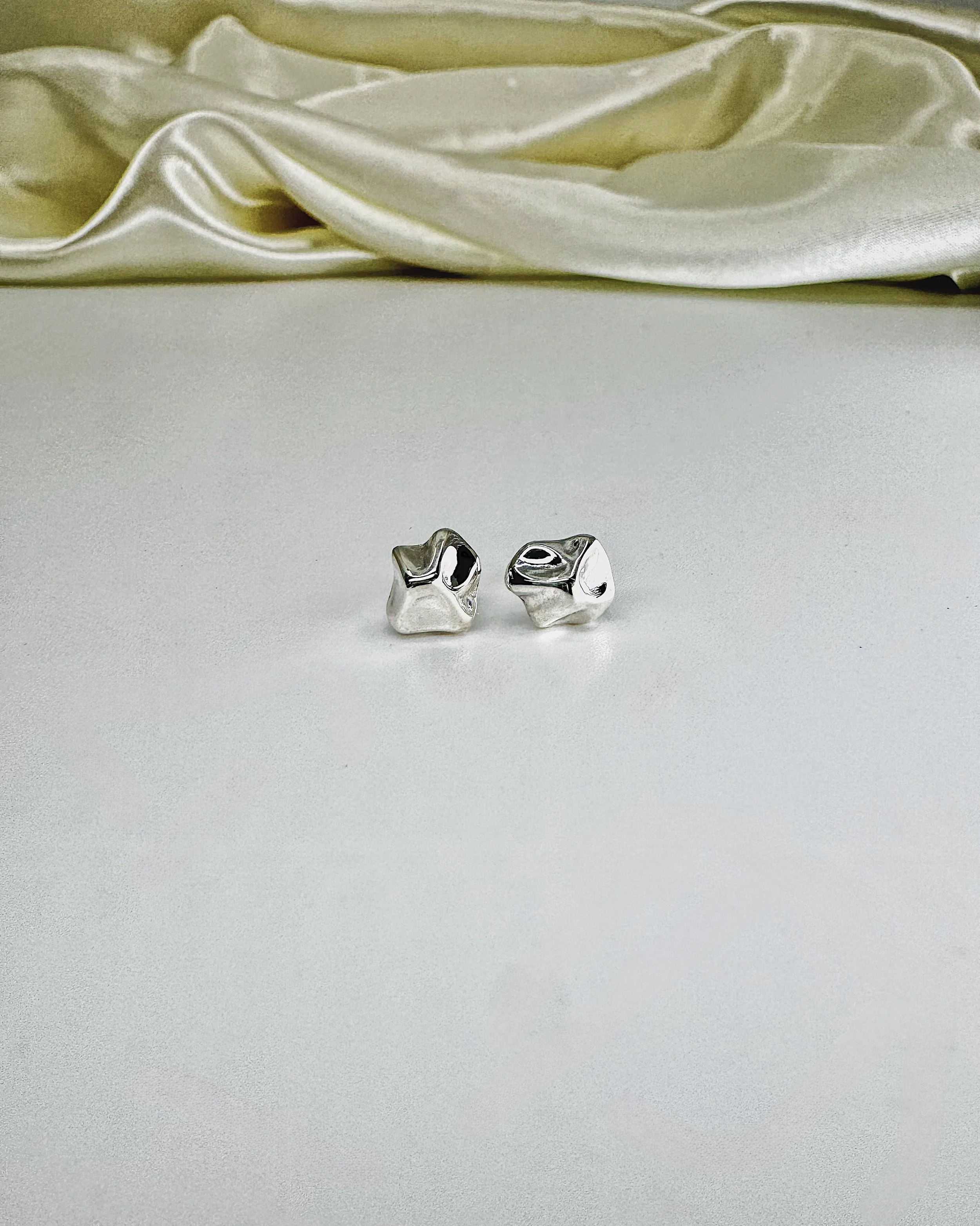 Two shiny silver miniature skulls placed on a smooth, light-colored surface with a textured pattern, and a satin fabric with folds in the background.