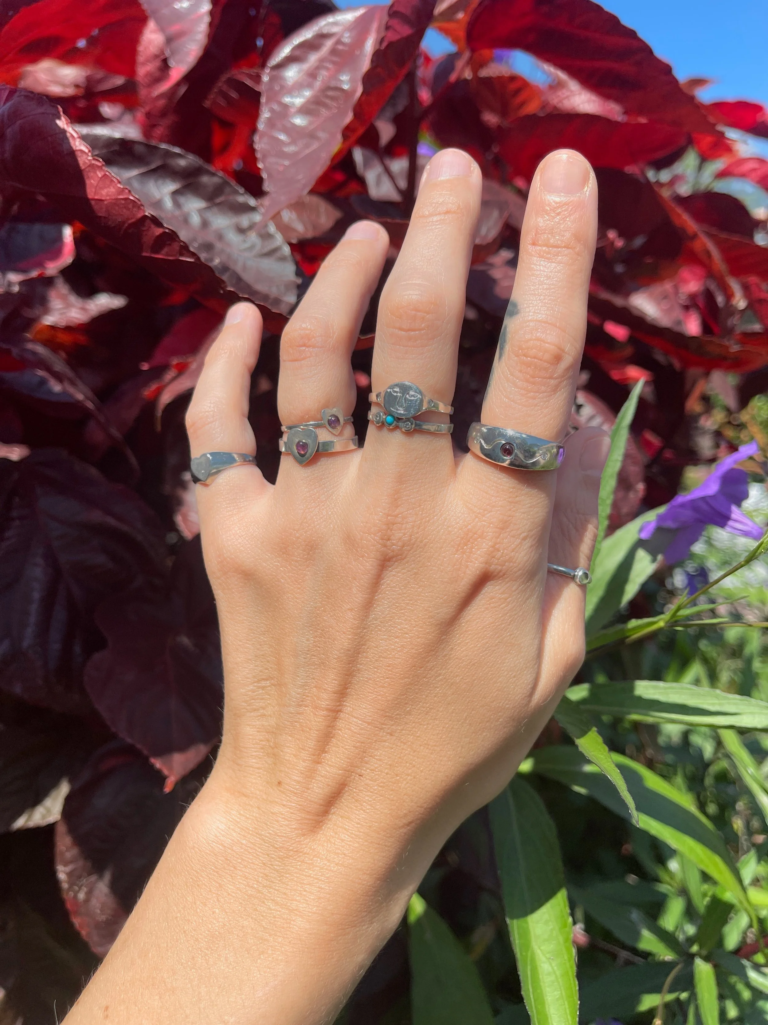 A hand with multiple rings displayed against a background of large, dark red and green leaves and purple flowers.